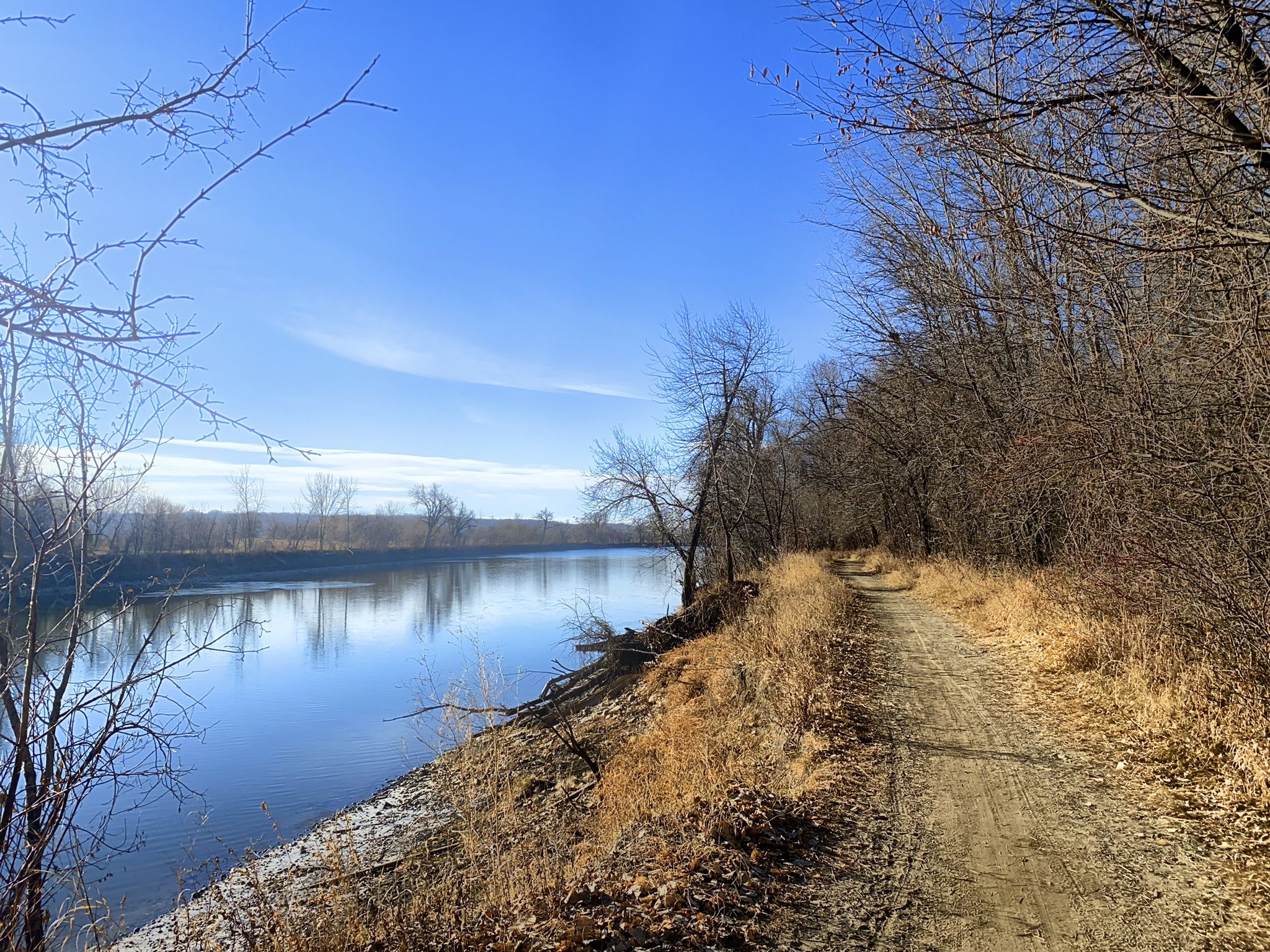Dirt path alongside a calm river, surrounded by bare trees under a bright blue sky. - Long Meadow Lake Trail