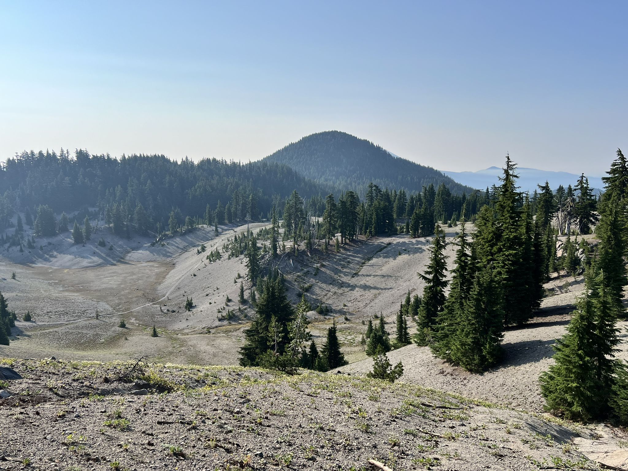 A wide valley with sparse trees, sandy slopes, and a distant forested mountain. - South Sister Climber Trail