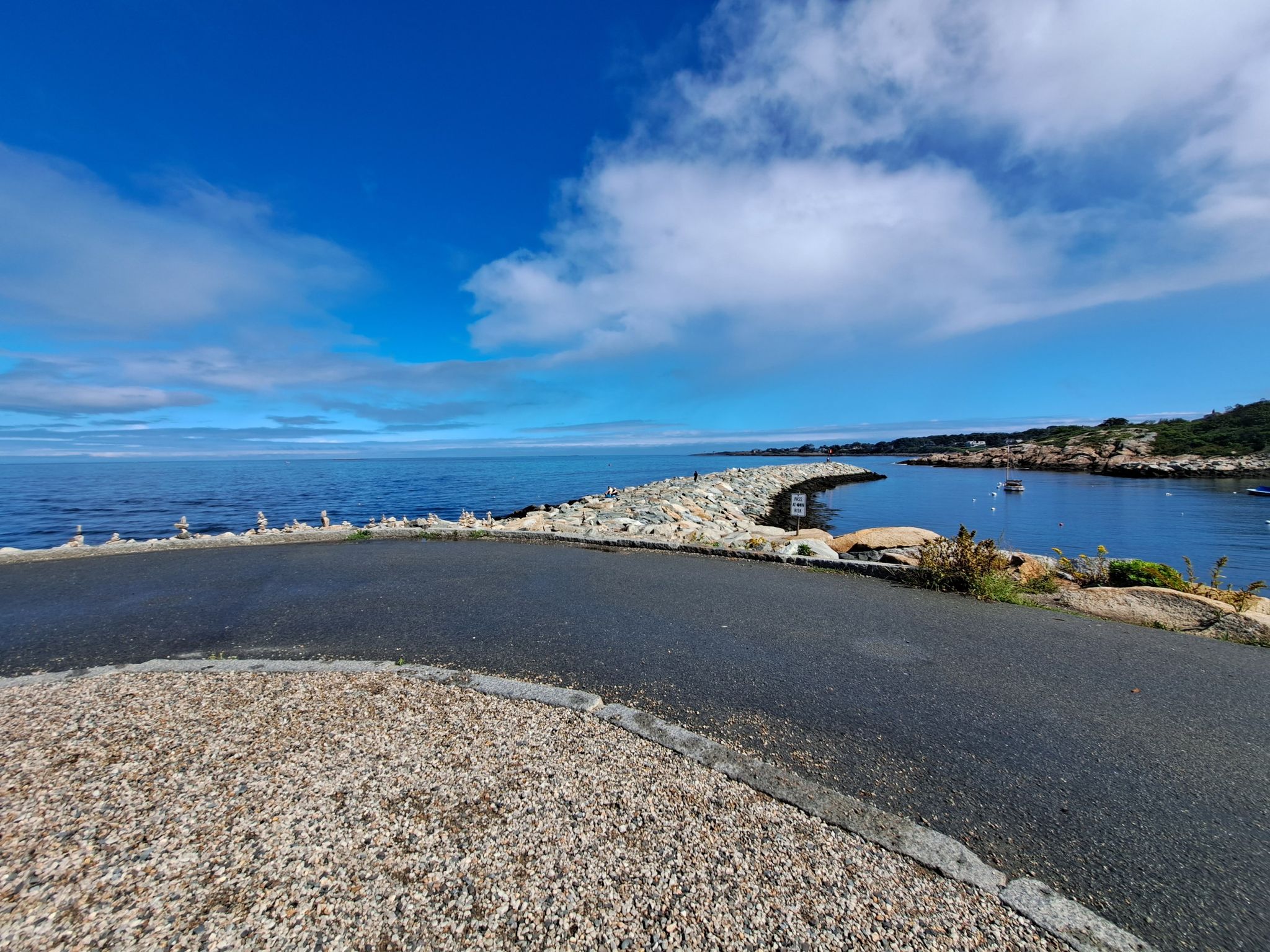 Paved road leads to a rocky breakwater under a bright blue sky. - Rockport Harbor – Bearskin Neck Lookout loop from Rockport