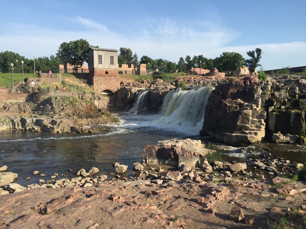 Sunny view of a wide waterfall cascading over rocks, with an old building nearby. - Falls Park loop