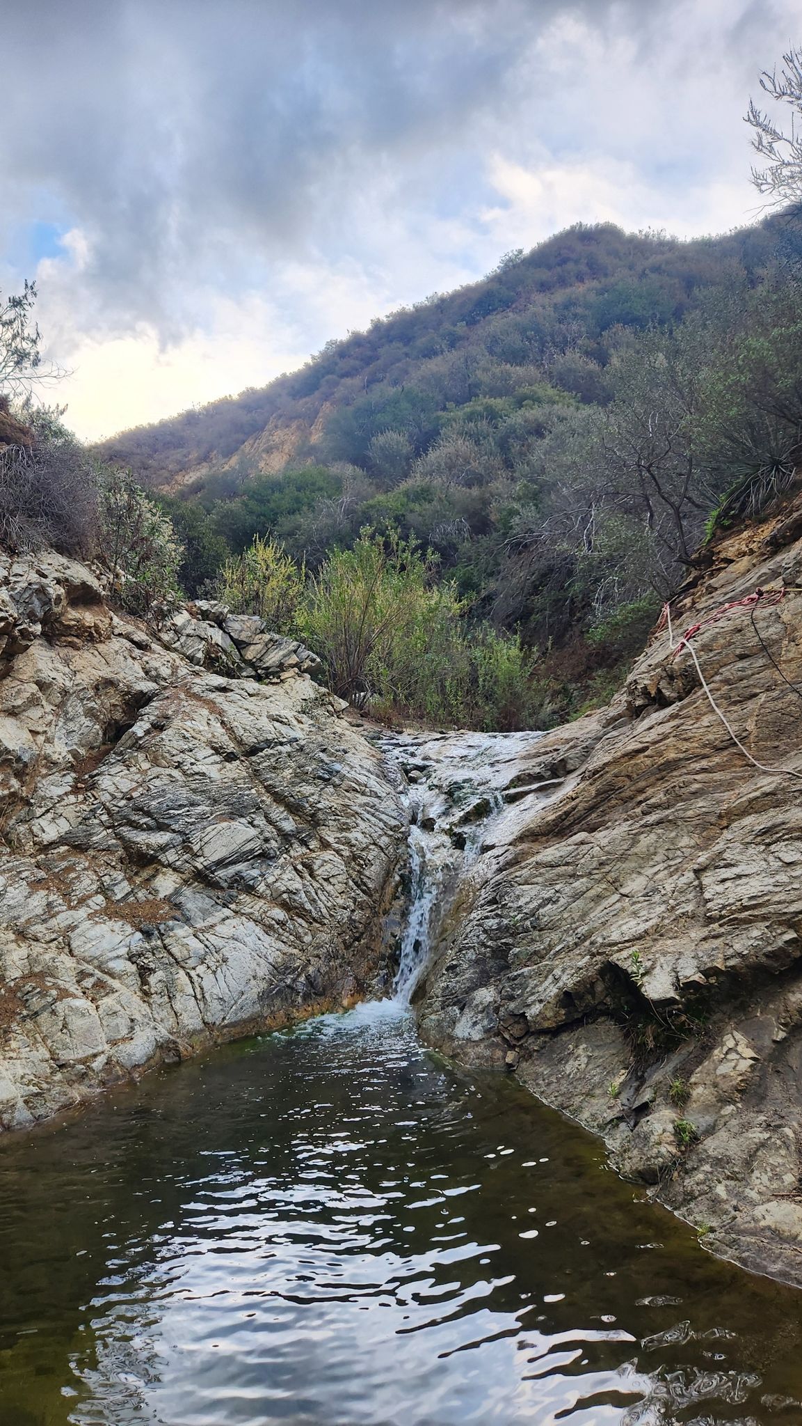Small waterfall cascades into a rocky pool under a cloudy sky. - Whitney Canyon Road Trail