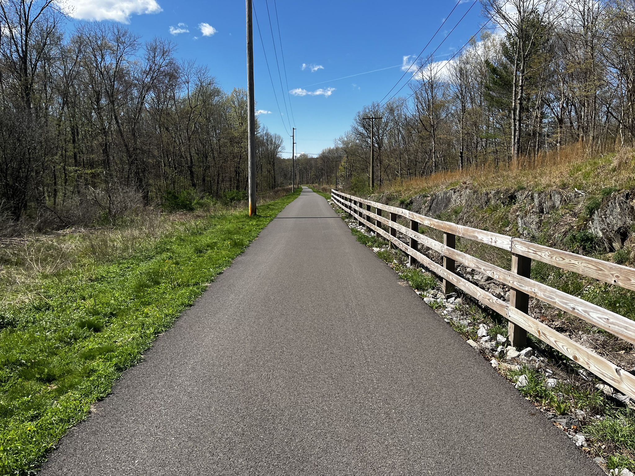 Paved path through a forest with a wooden fence under a bright blue sky. - Nassau Lake – Albany-Hudson Electric Trail loop from Village of Nassau