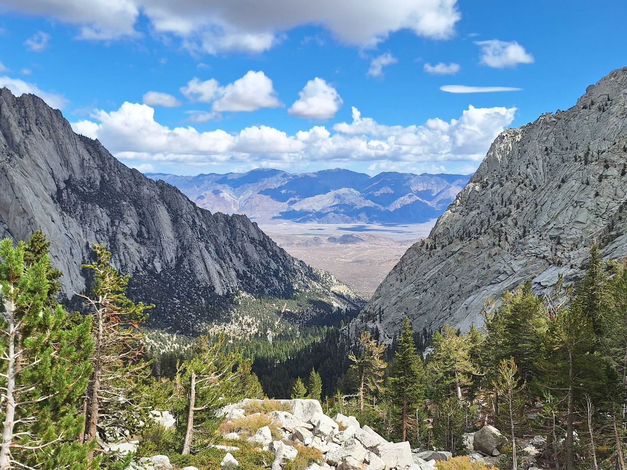 Mountain valley with pine trees, distant desert landscape, and cloudy blue sky. - Mount Whitney Trail
