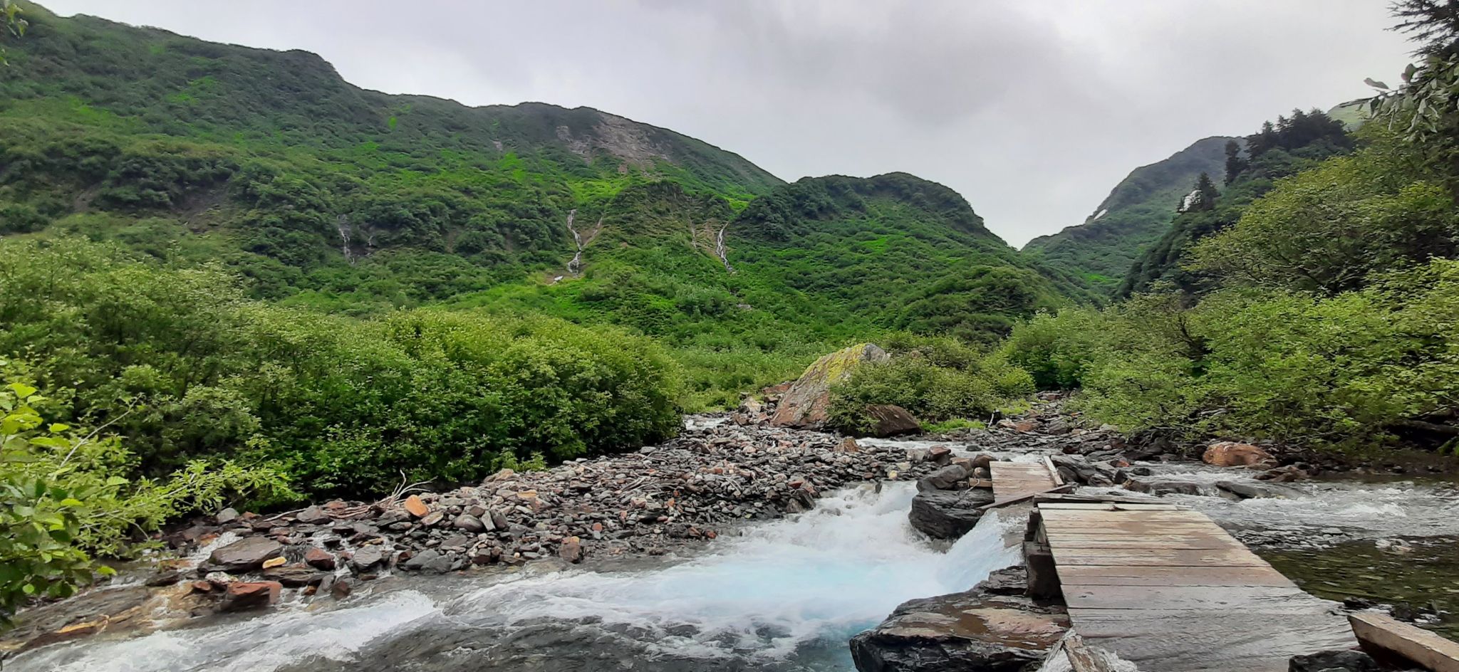 Rushing river with a wooden walkway, surrounded by lush green mountains under cloudy skies. - Perseverance Trail
