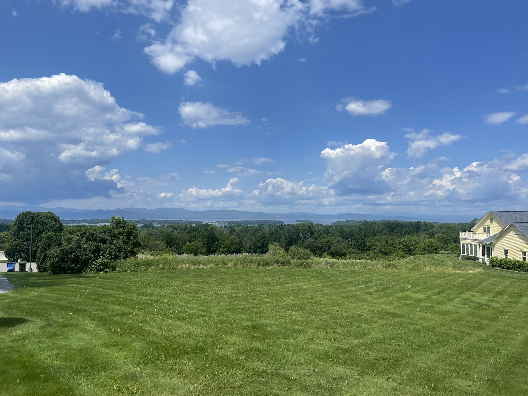 Bright sunny day over a green field, distant mountains, and a house. - Winooski River to Shelburne Pond loop