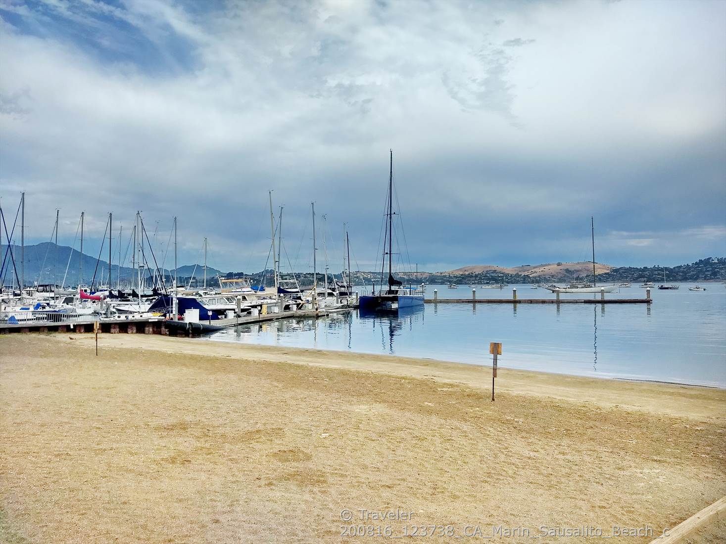 A sandy beach next to a marina with many sailboats under a cloudy sky. - Schoonmaker Beach – Dunphy Park loop from Sausalito