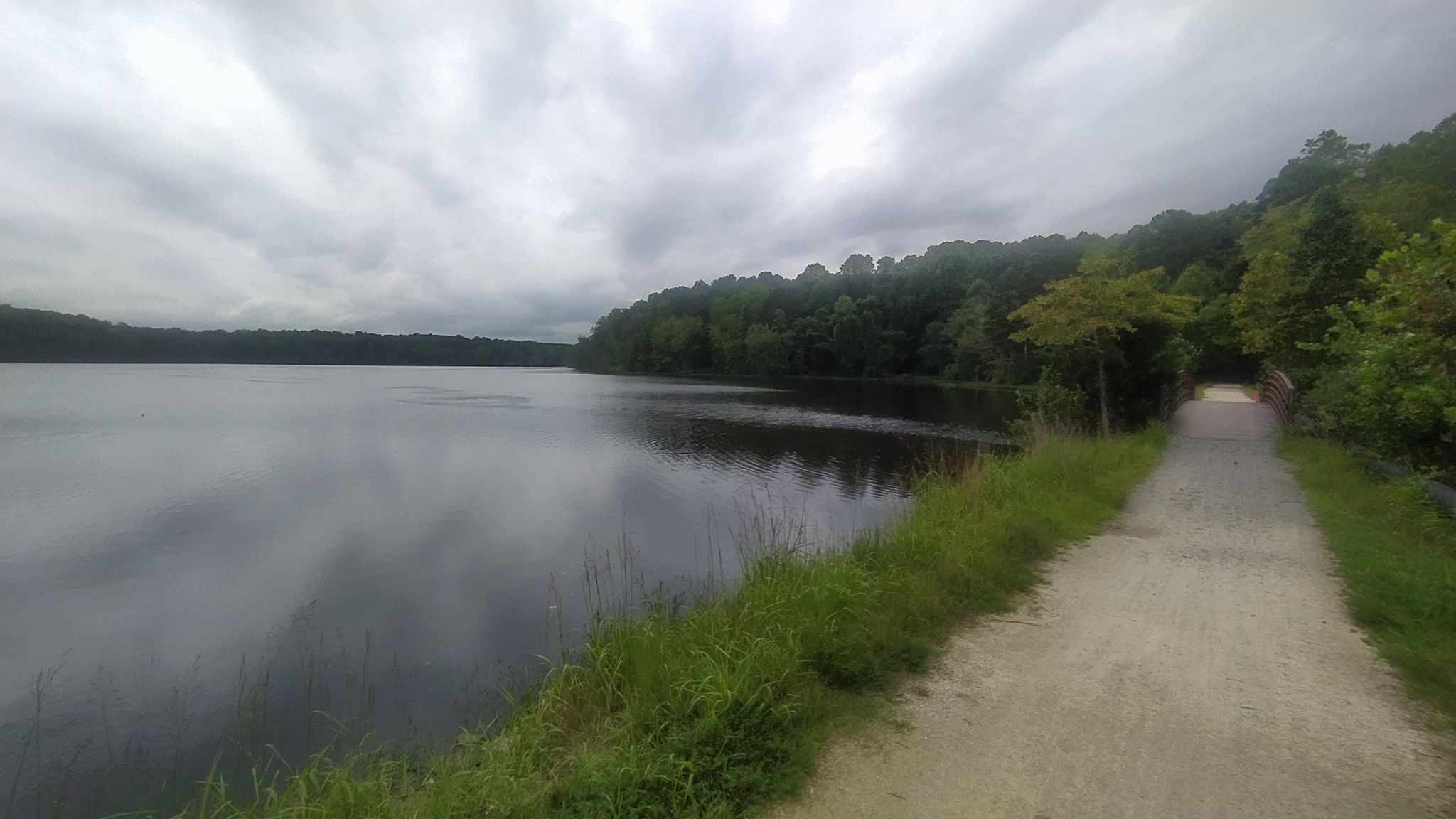 A dirt path runs alongside a calm lake under an overcast sky. - Cascades Preserve gravel loop via Quarry Road
