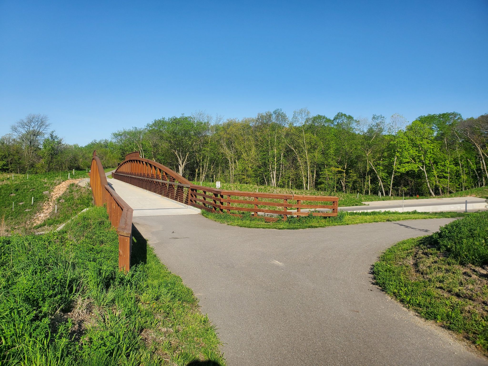 Paved path forks before a wooden arched bridge, surrounded by green trees. - Minnesota River Bluffs LRT Regional Trail
