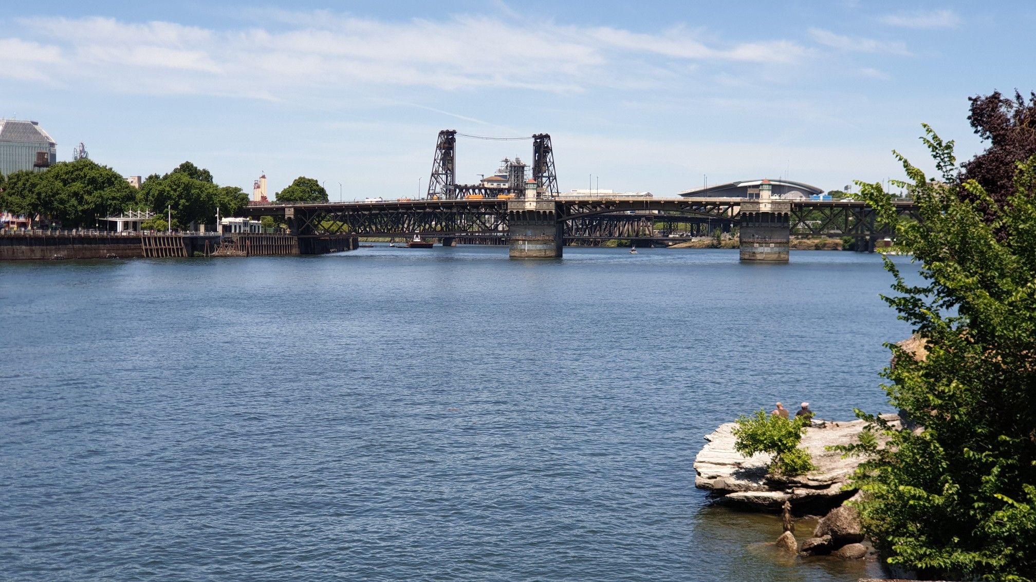 A large, dark metal bridge spans a wide river on a sunny day. - Eastbank Esplanade and Waterfront Park Loop