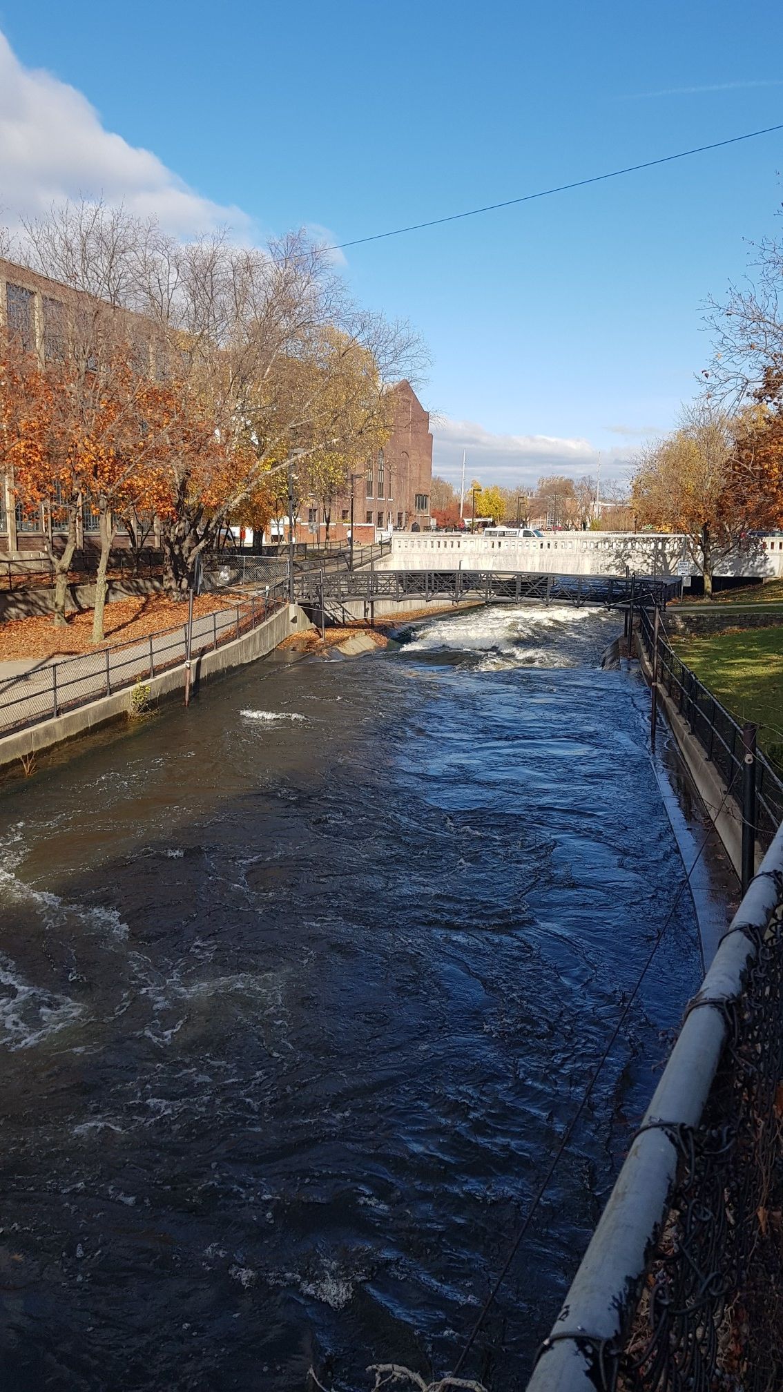 Narrow river with rapids flowing past urban buildings and autumn trees. - East Race Waterway – St Joseph River bridge loop from James Whitcomb Riley High School