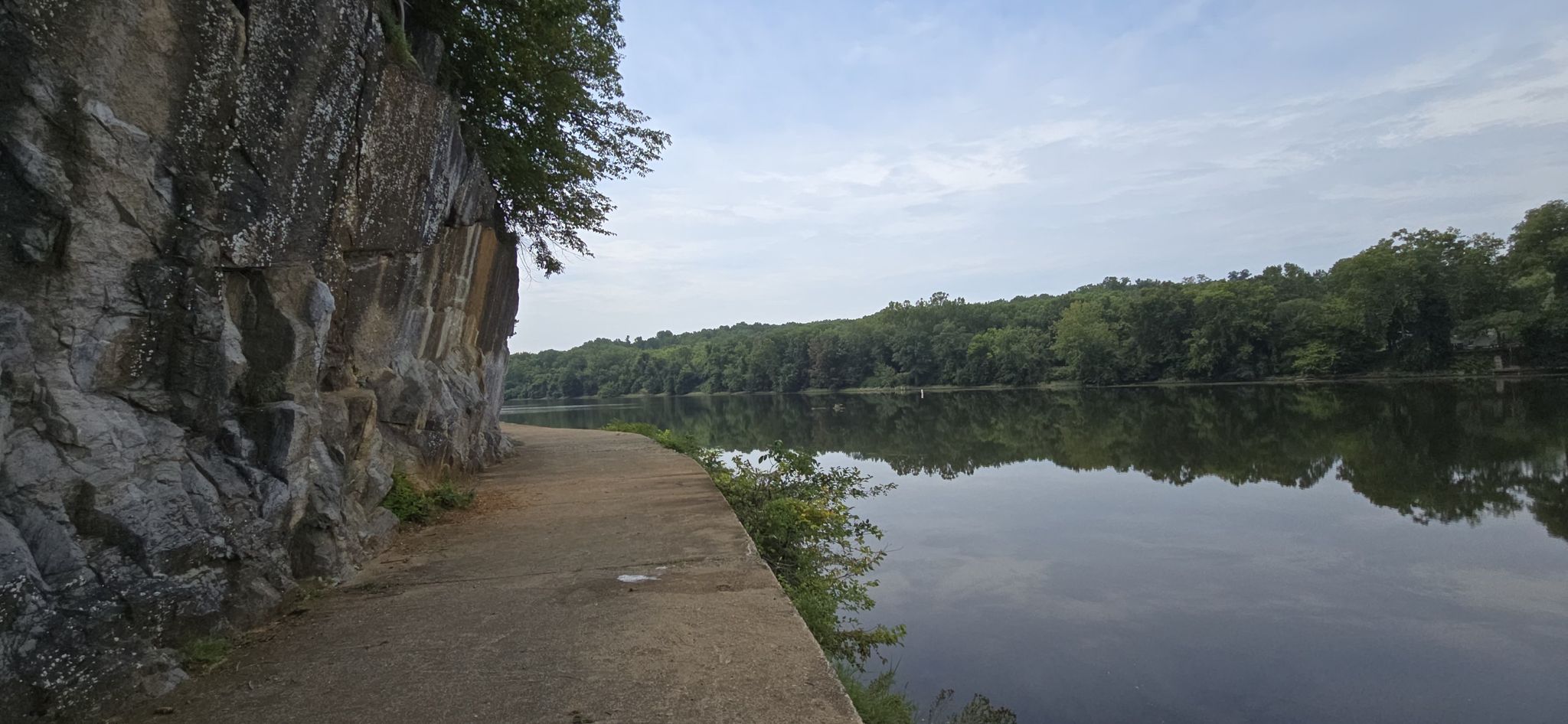 A paved path runs alongside a calm river, with a rocky cliff and trees. - Dam Number 5 – C & O Bicycle, Hancock loop from Williamsport