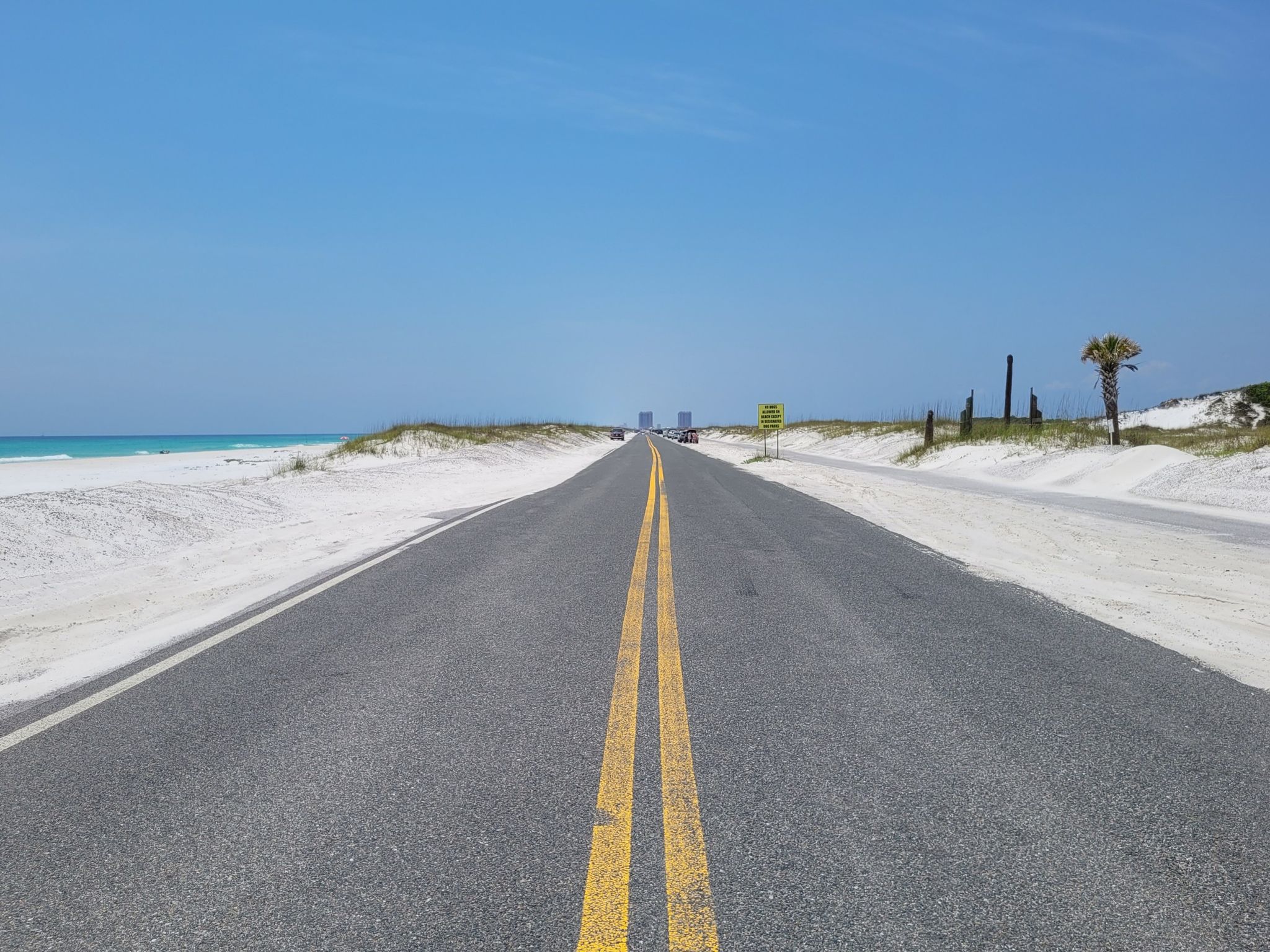 Paved road with yellow lines, flanked by white sand dunes and ocean. - Park East On-Leash Dog Beach – Sugar Bowl Protected Area loop from Pensacola Beach