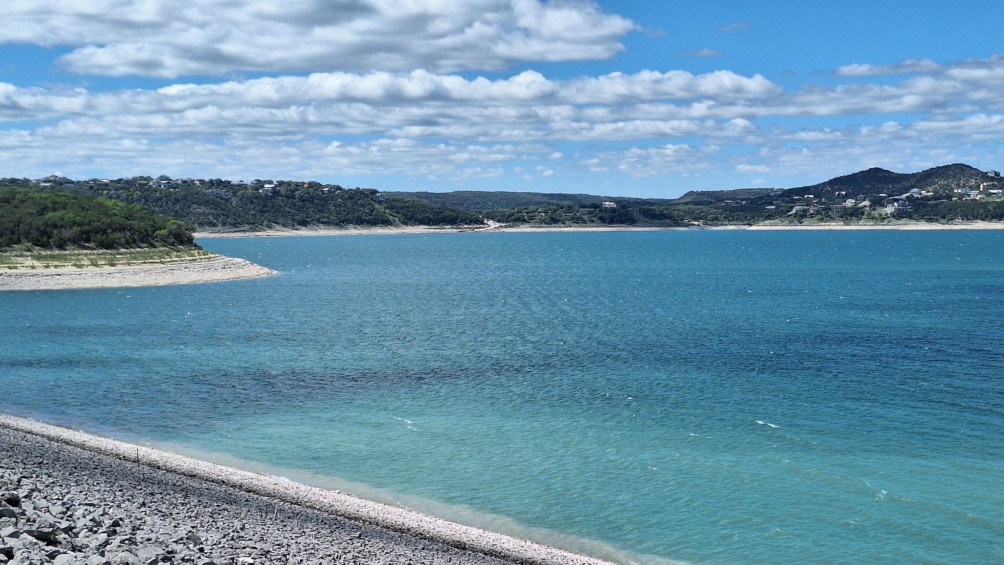 Bright blue water, rocky shoreline, and distant hills under a sunny, cloudy sky. - Dam Crest Trail