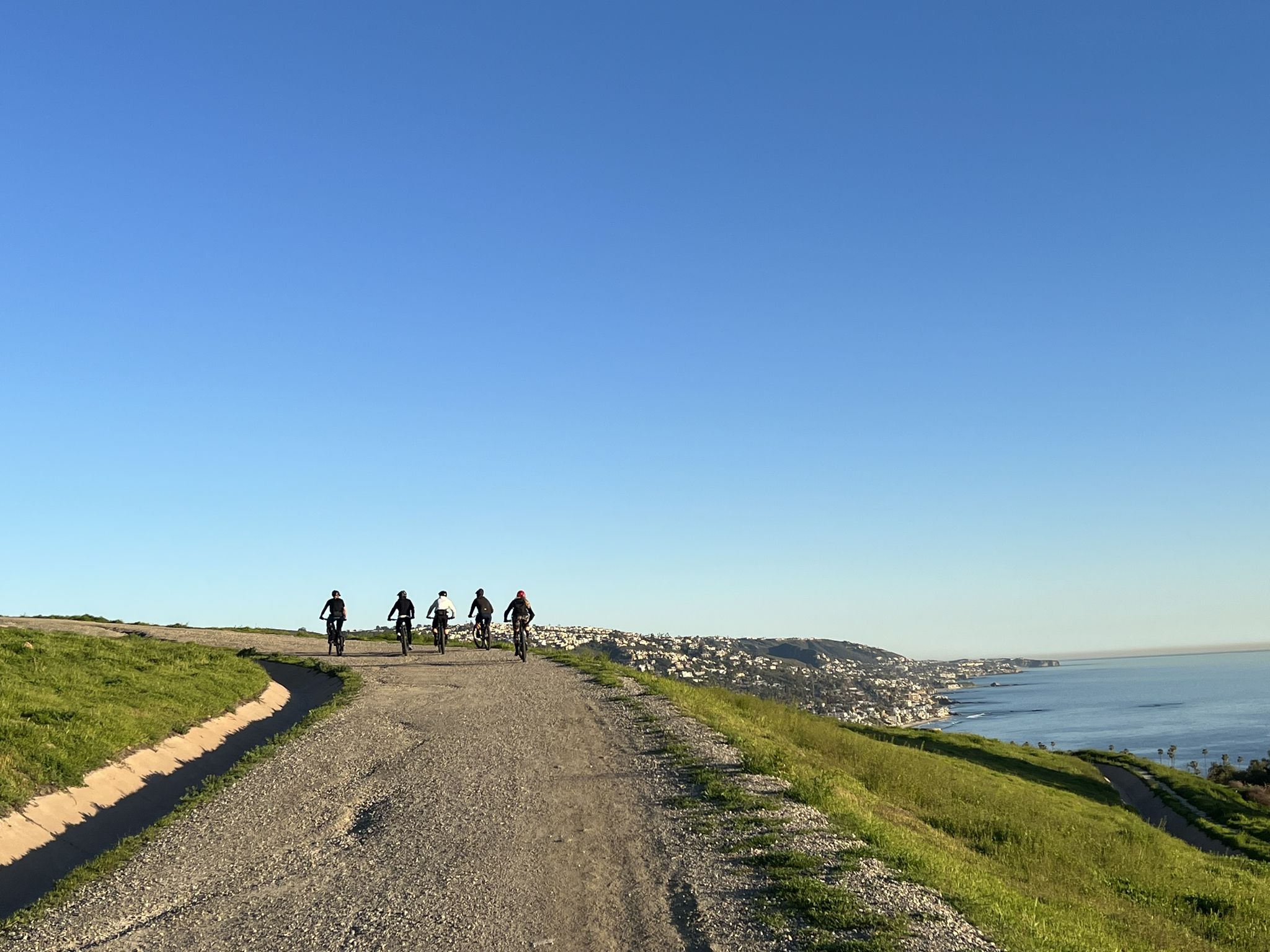 Five cyclists ride a dirt path on a grassy hill overlooking the ocean. - Bommer Ridge – Boat Road loop from Laguna Beach