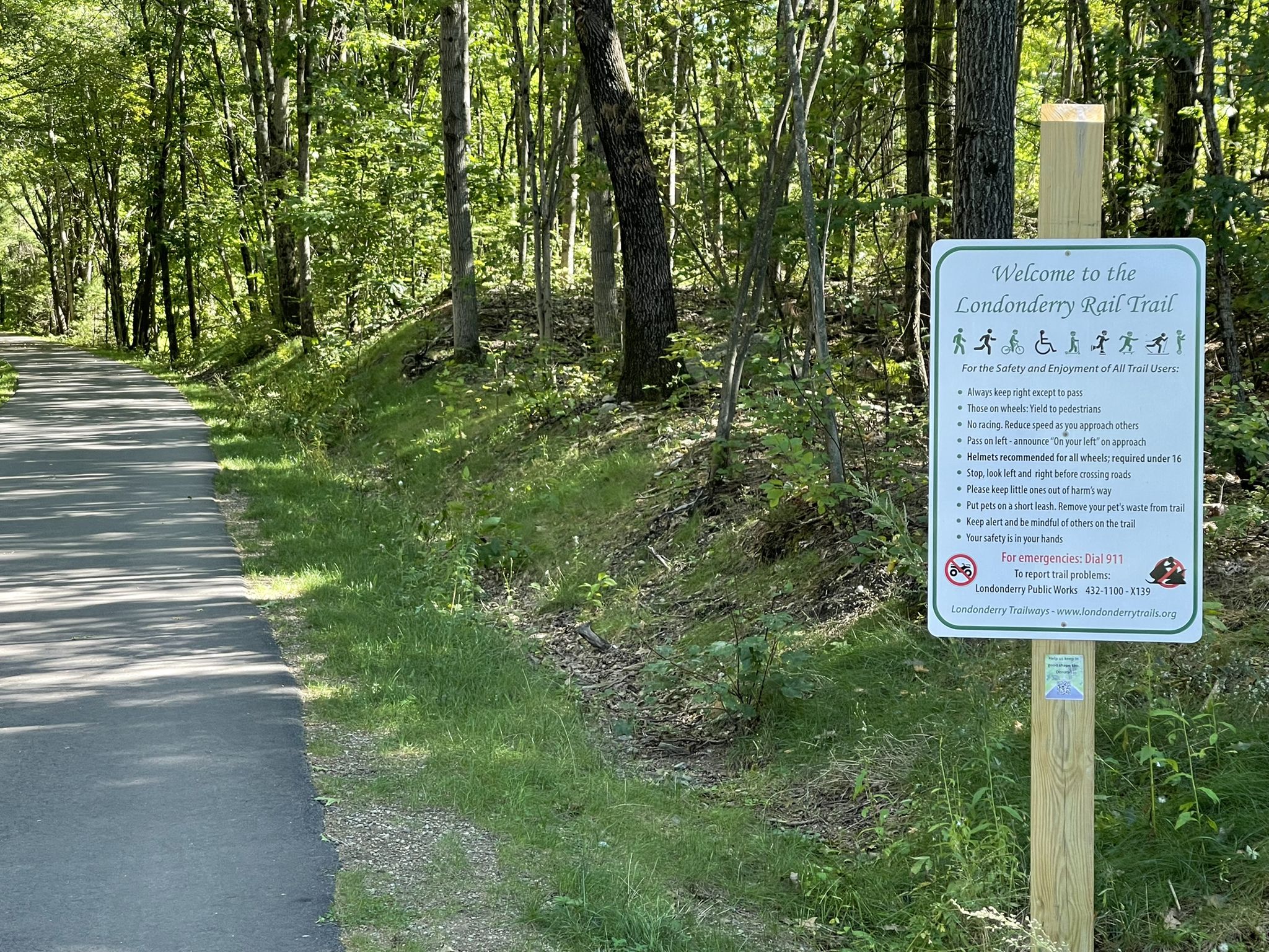 Paved path through a sunny forest with a trail information sign. - Londonderry Rail Trail – Londonderry Rail Trail loop from Wilson