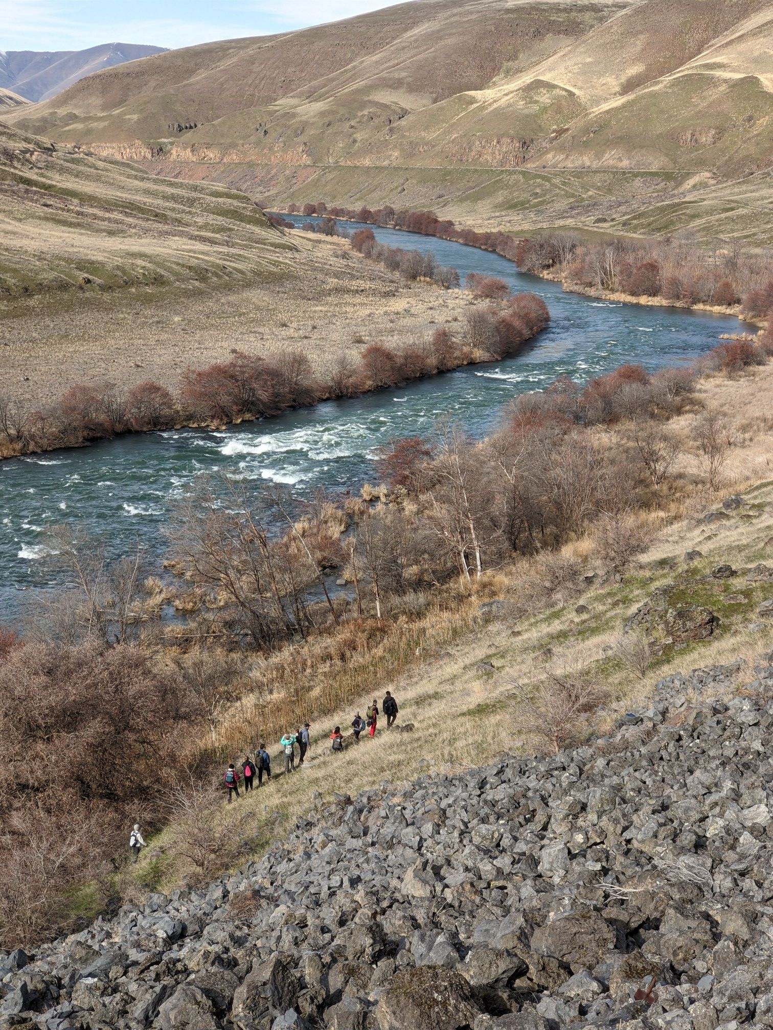 A group of people hike down a rocky slope overlooking a winding river. - Deschutes River Trail