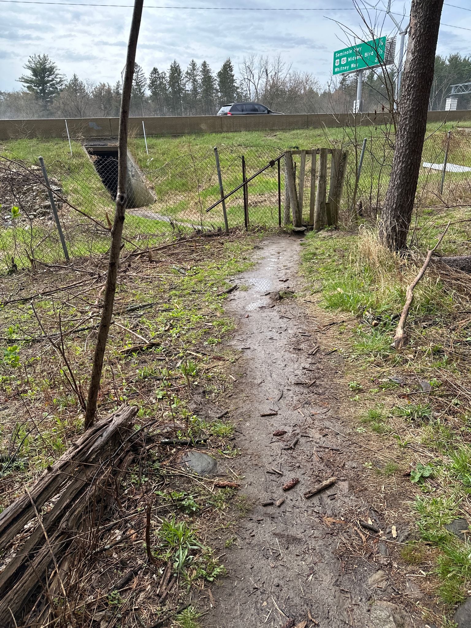 A narrow dirt path leads to a wooden gate near a highway overpass. - Lake Wingra loop from Madison
