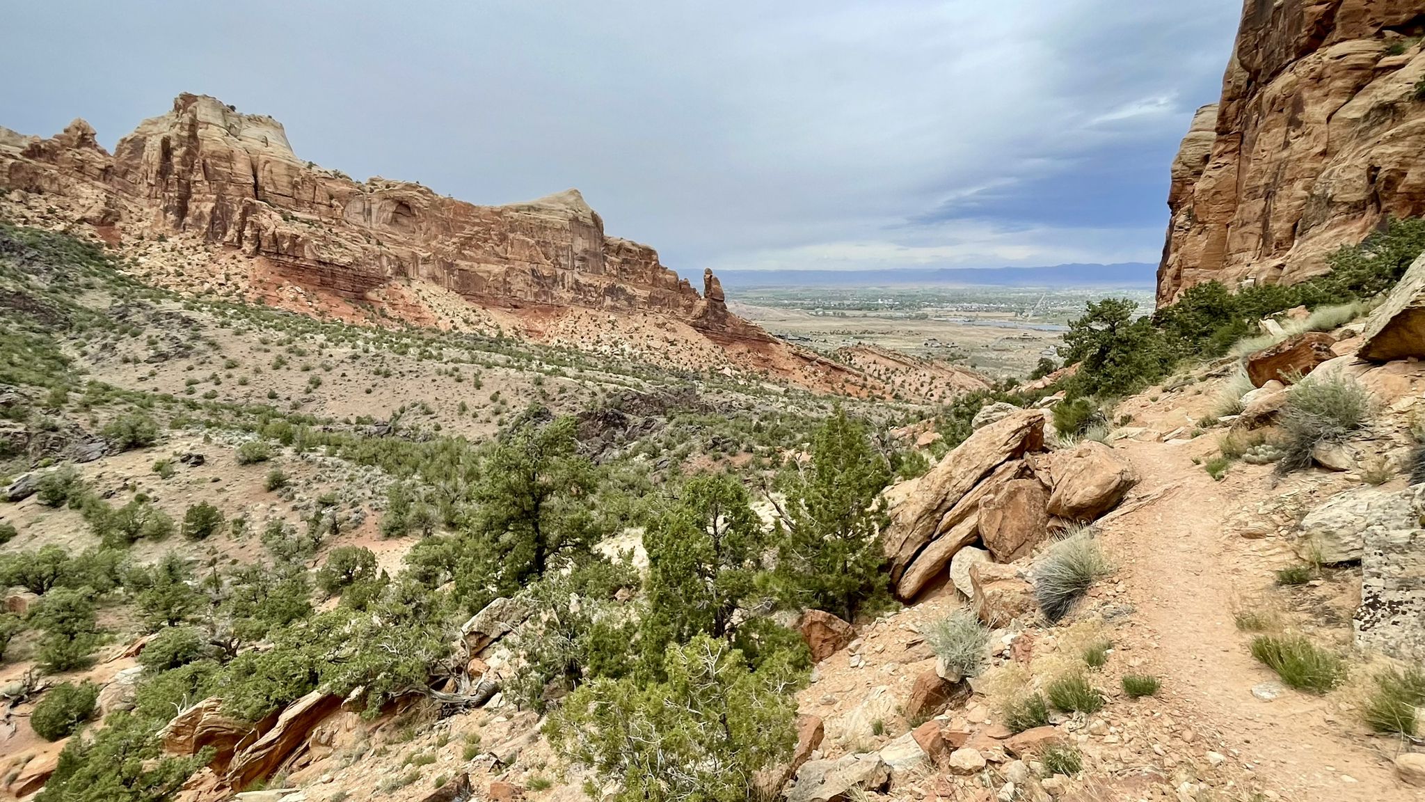 A dirt trail overlooks a vast desert valley with distant mesas under a cloudy sky. - Monument and Wedding Canyon Loop