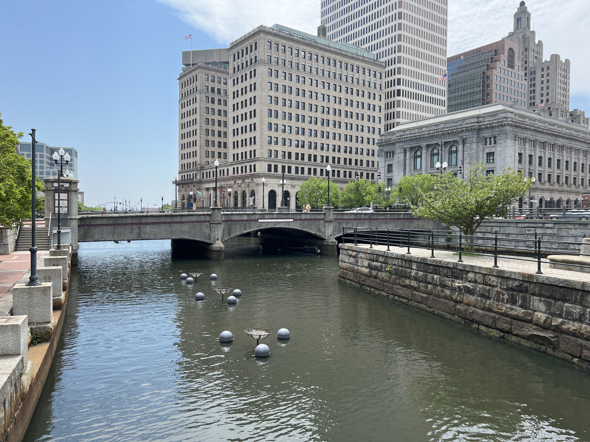 Urban river with a bridge, tall buildings, and floating spheres on a sunny day. - East Coast Greenway: Providence Loop