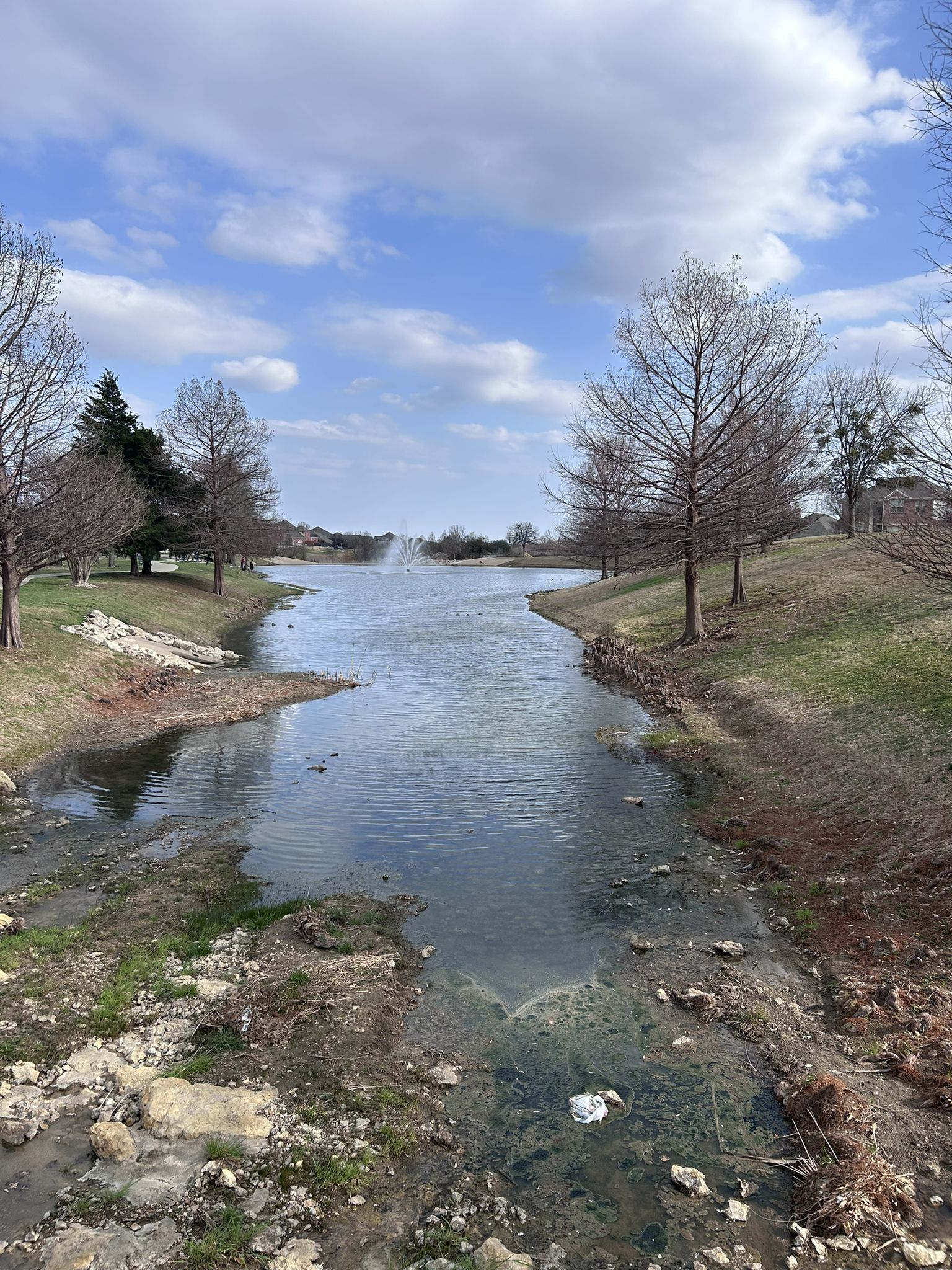Narrow pond with a distant fountain, bare trees, and green banks under cloudy sky. - Crawford Farms Park & Sunset Hills Park loop