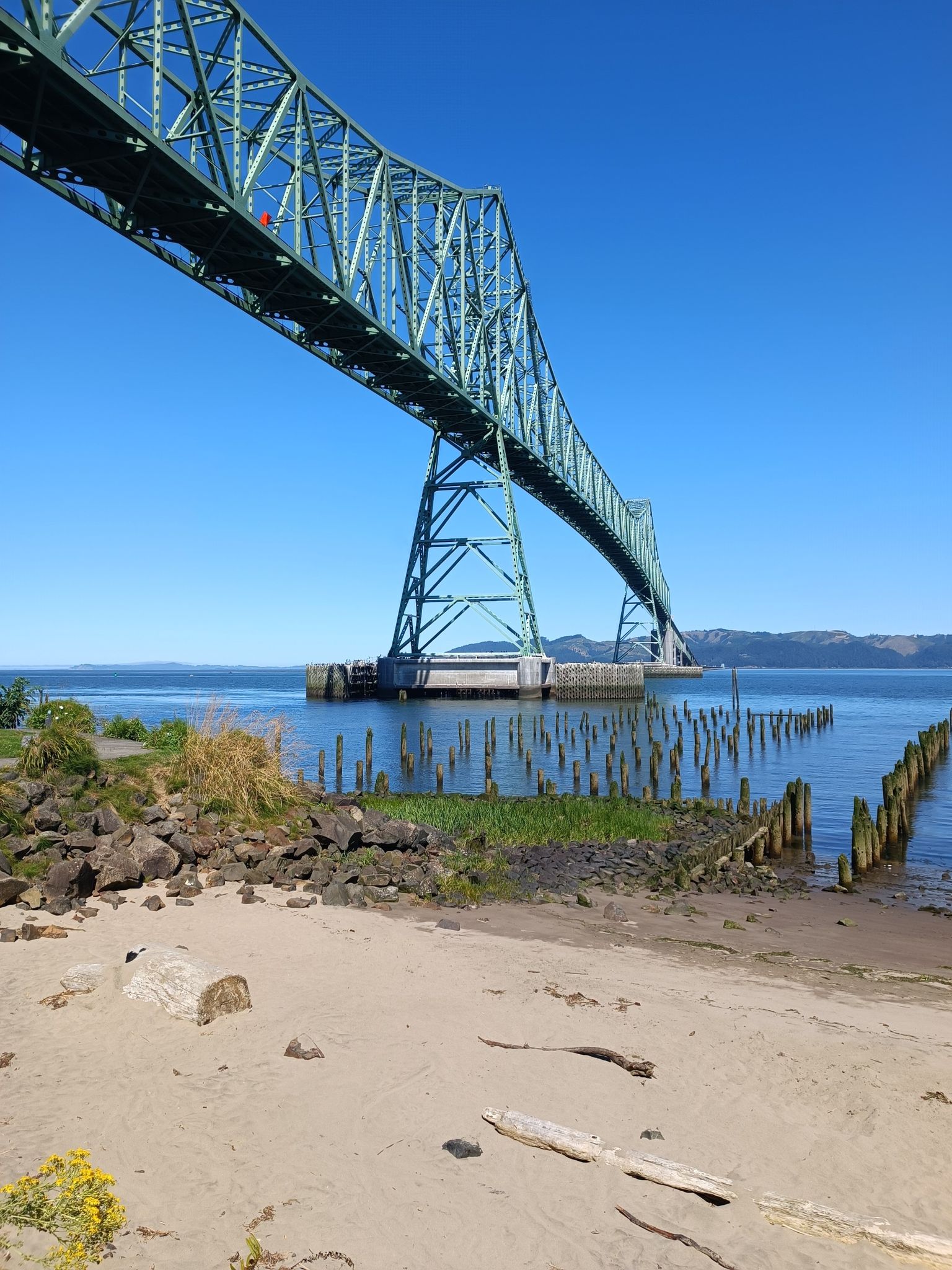 Green steel bridge over sandy beach and water, clear blue sky. - Astoria Riverwalk Trail