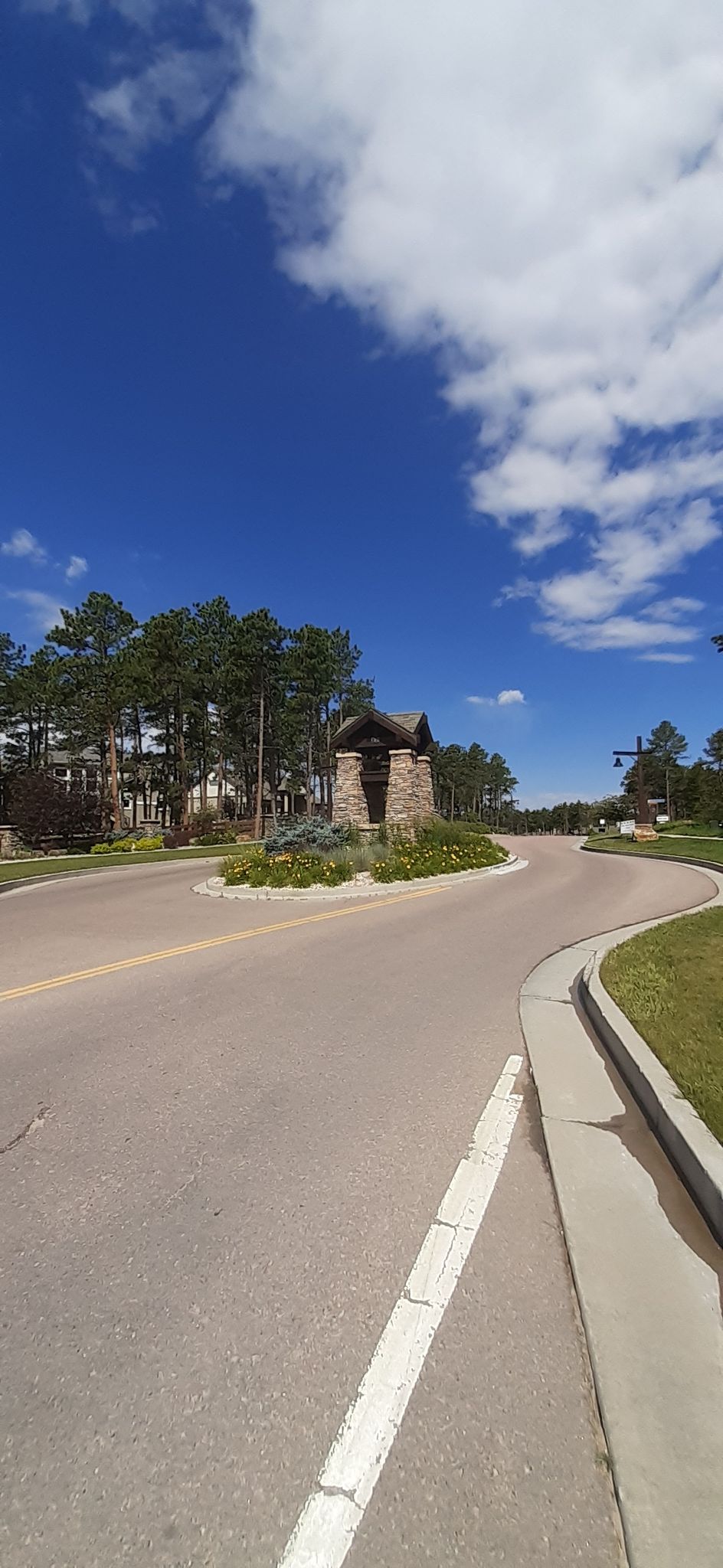 Sunny day, road roundabout with stone structure, tall trees, and blue sky. - Fox Run Regional Park loop