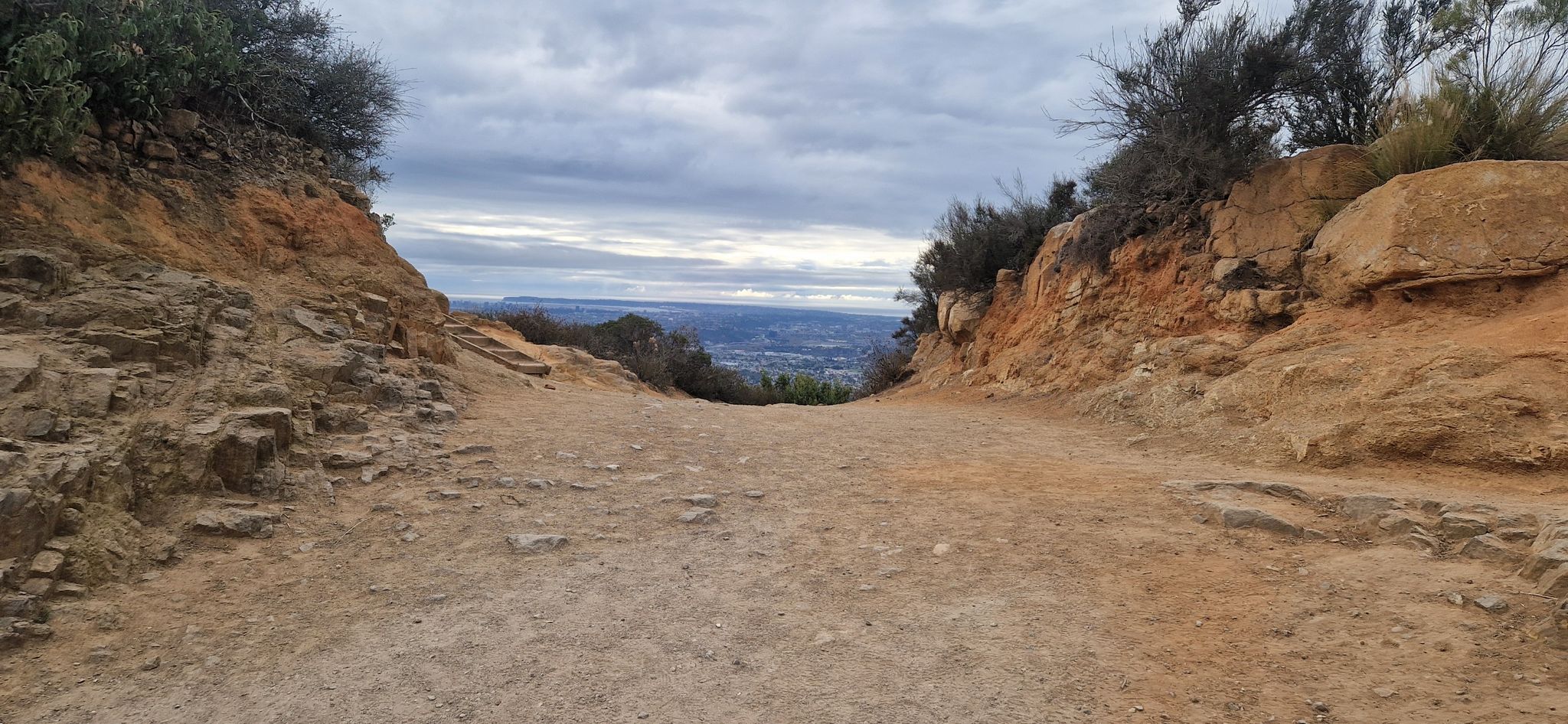 Dirt path through rocky cuts, leading to a distant hazy city. - Big Rock and Mesa Trail Loop