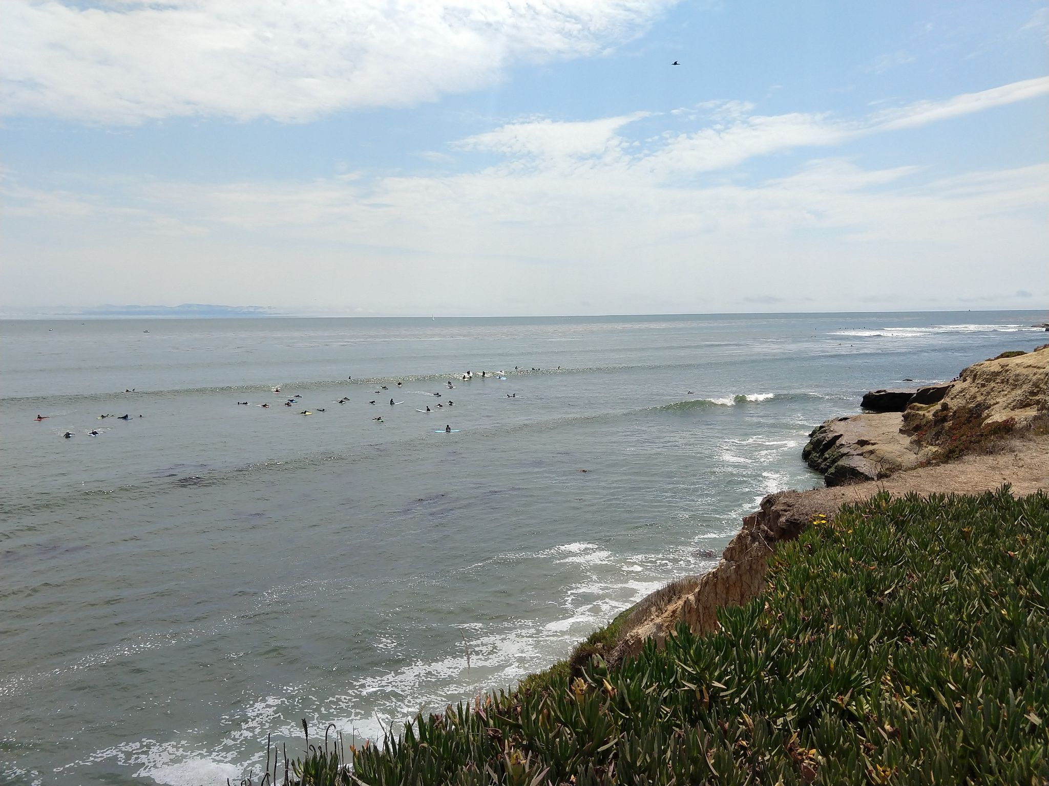 Ocean with many surfers, waves, and a rocky cliff under a bright sky. - East Cliff Drive County Parkway