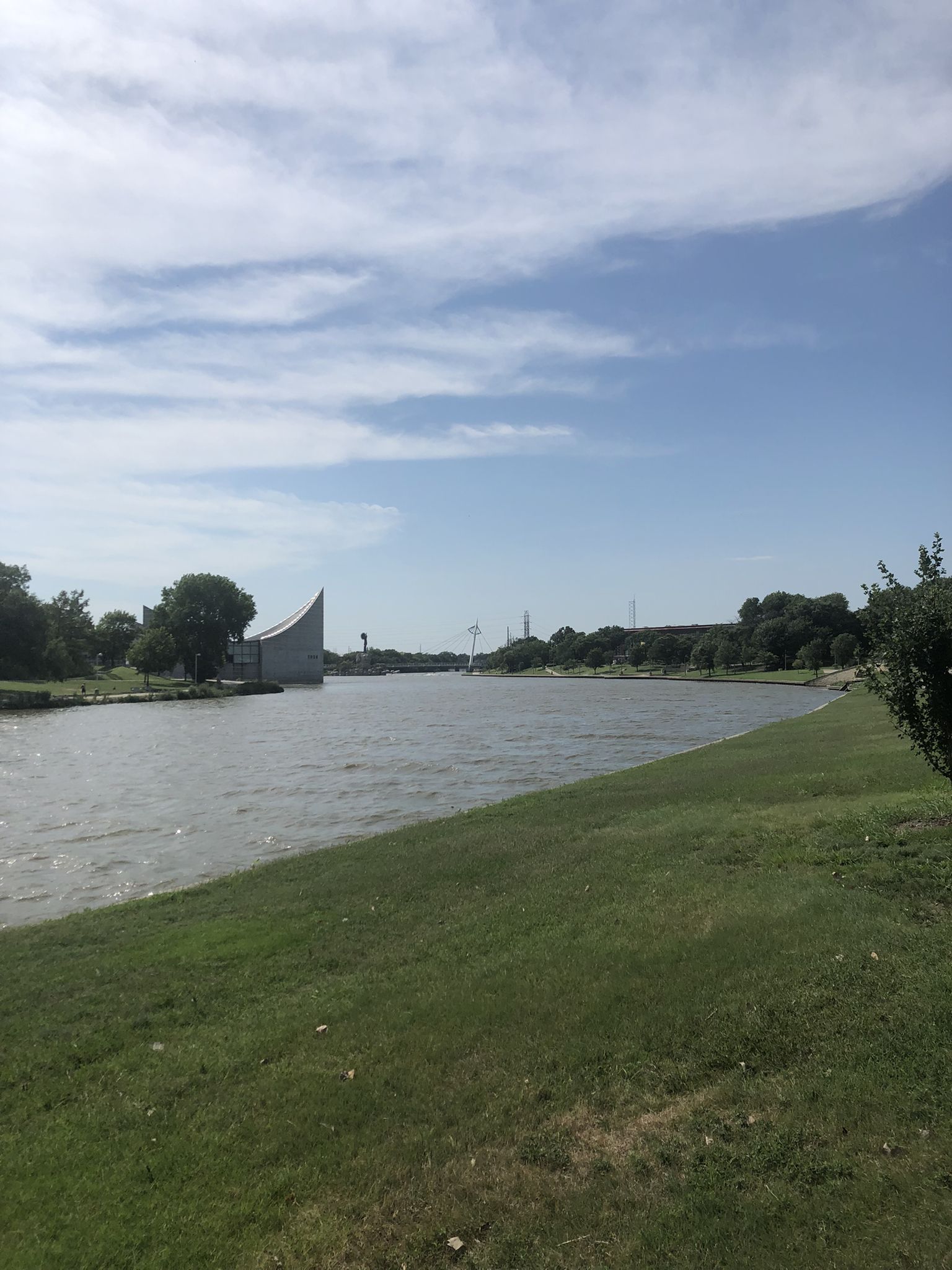 Wide river with a green bank and distant buildings under a bright, cloudy sky. - Keeper of the Plains Statue – Keeper of the Plains loop from Wichita