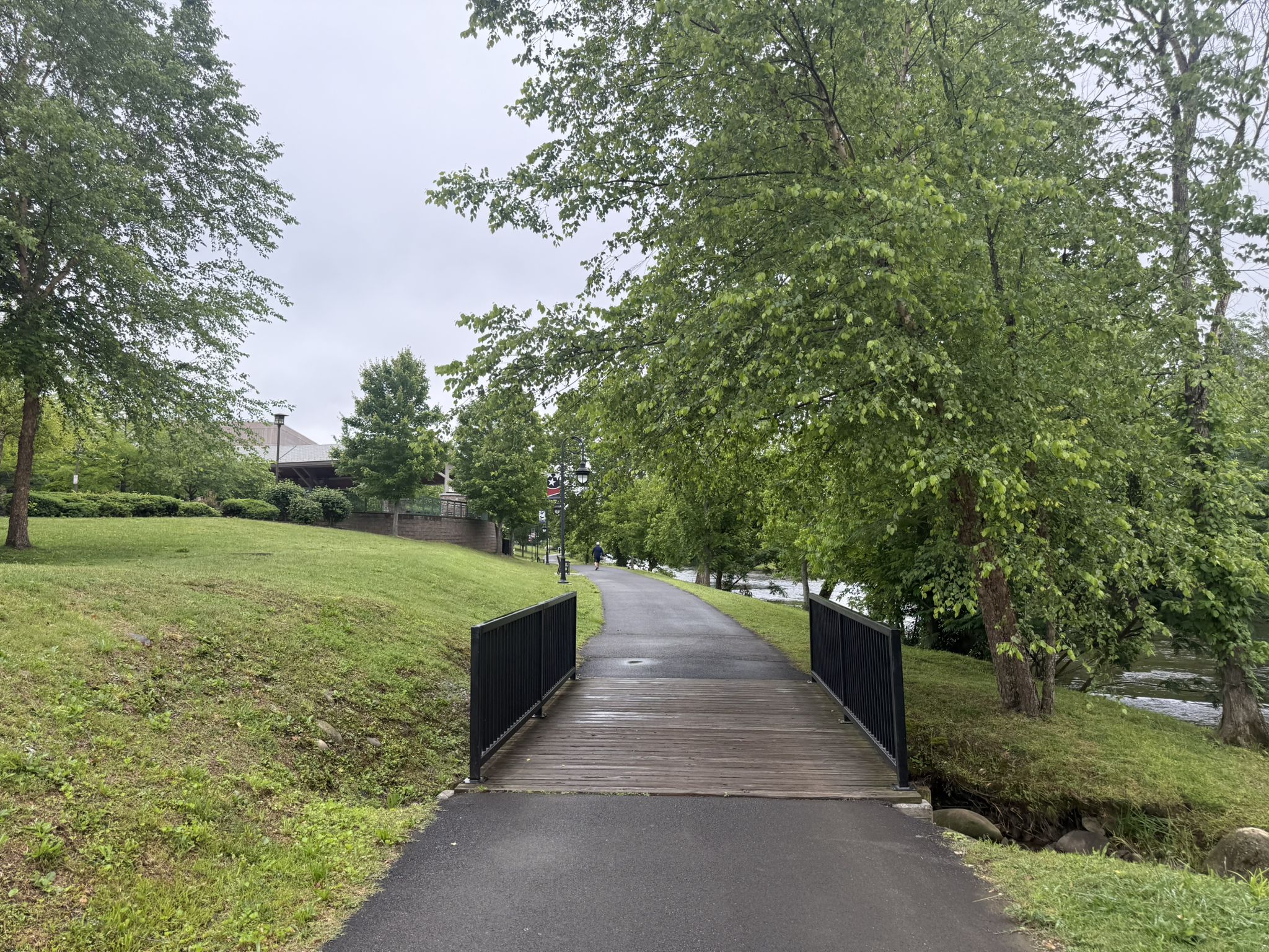 Paved path leading to a wooden bridge surrounded by green trees on an overcast day. - Pigeon Forge Riverwalk