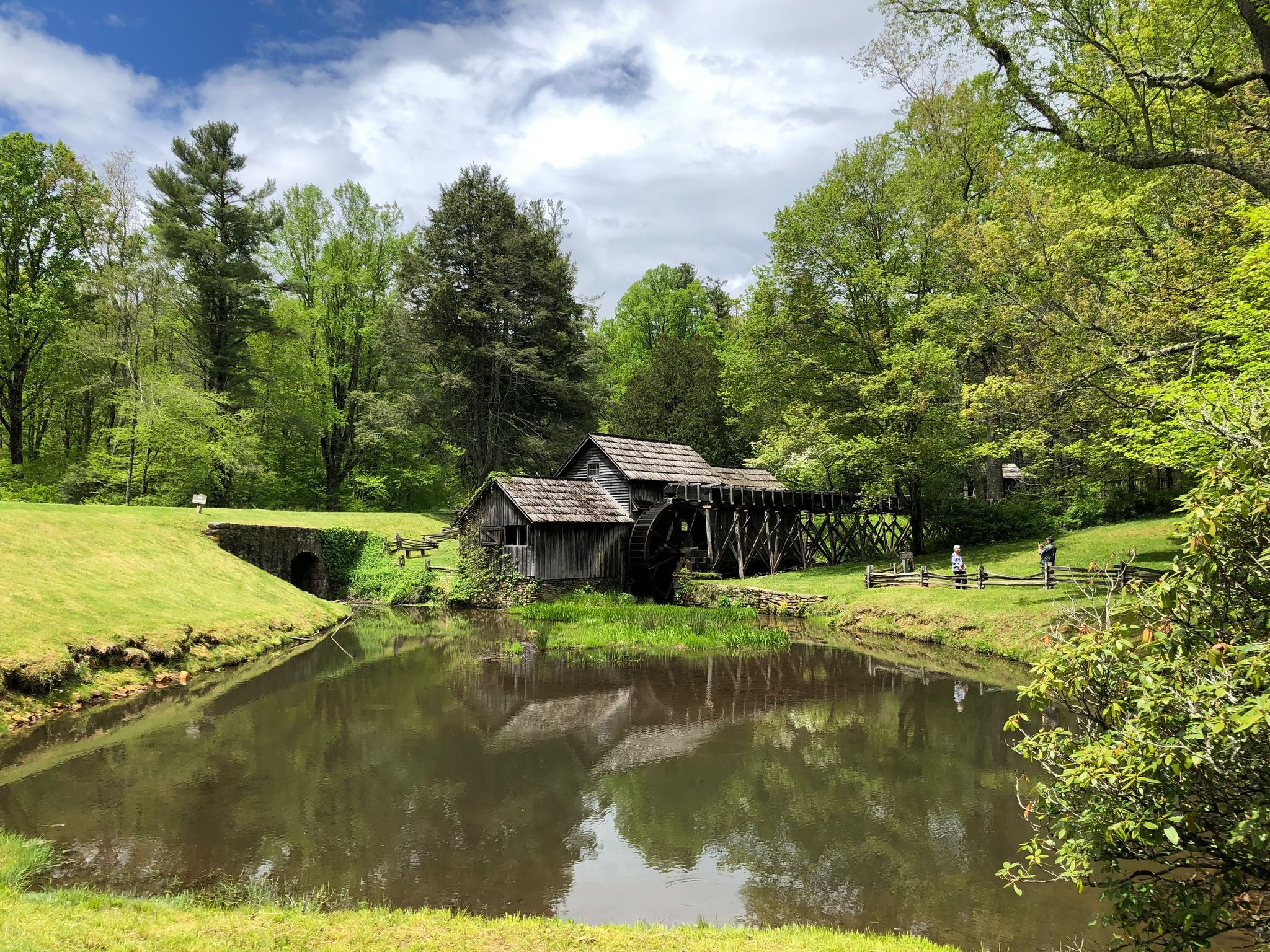 Old wooden mill by a reflective pond, surrounded by lush green trees under a bright sky. - Mountain Industry Trail