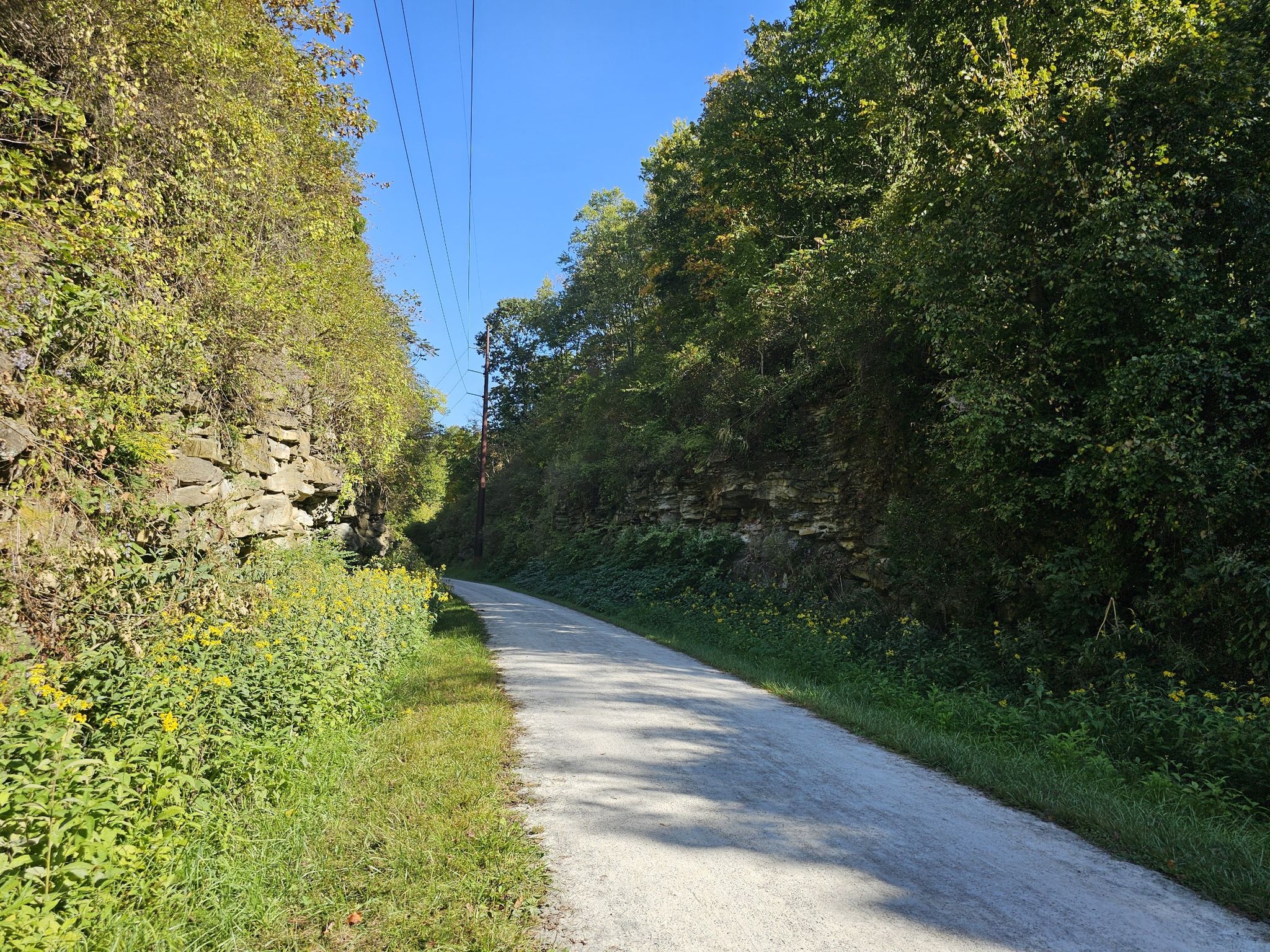 A gravel path between a stone wall and green trees under clear blue sky. - Bike loop from Glenfield
