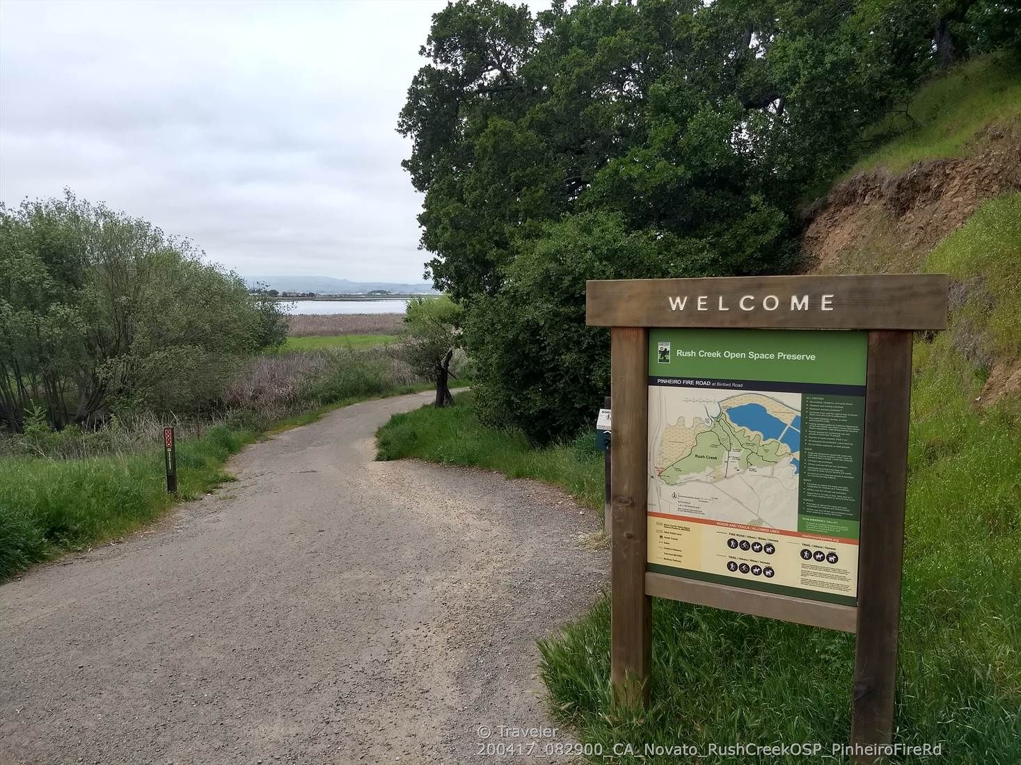 A welcome sign stands beside a dirt path, with distant water and trees. - 101 Trail