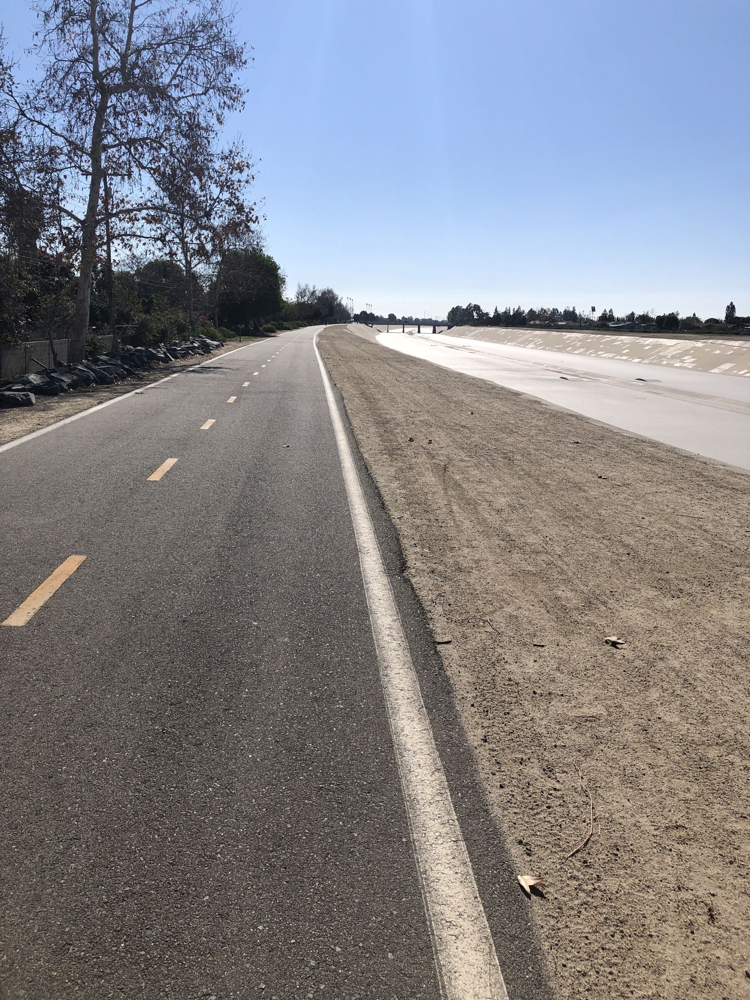 Sunny view of a paved road beside a wide, dry concrete channel. - Santa Ana River Trail loop from Santa Ana College