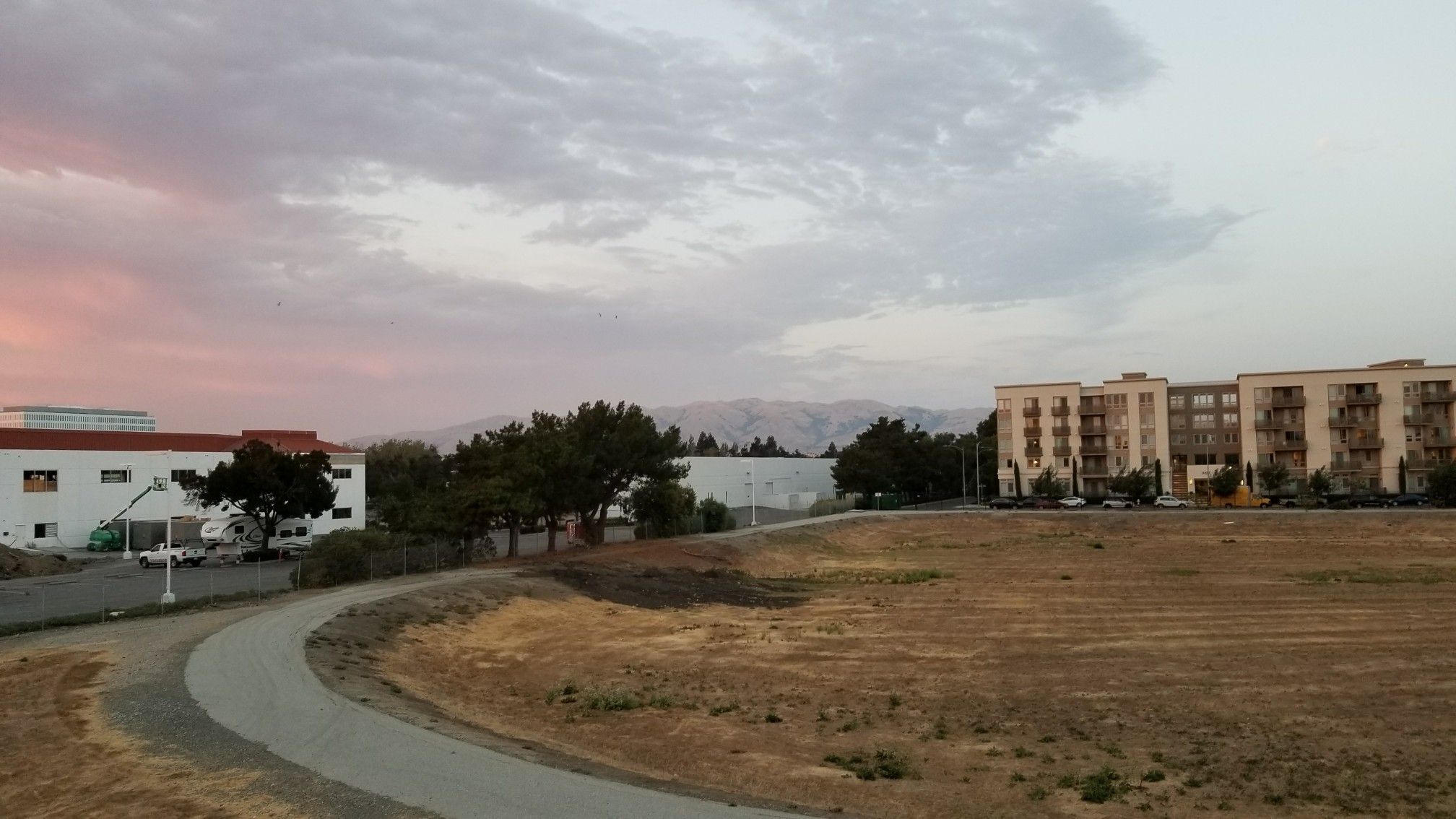 Curved paved path with buildings and mountains under a colorful sunset sky. - Guadalupe River Trail