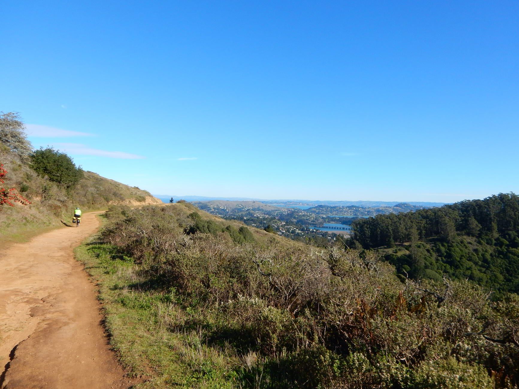 A person cycles on a sunny dirt path overlooking distant hills and water. - Tennessee Valley Trailhead – Panoramic Highway loop from Golden Gate National Recreation Area