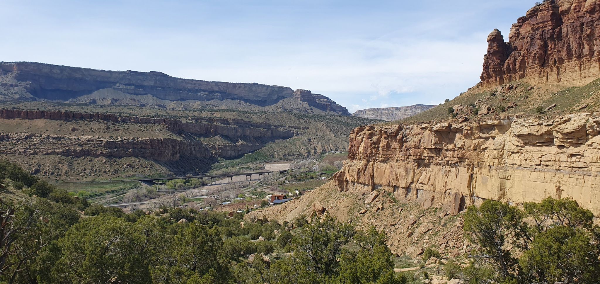 Bright day over a wide canyon with green vegetation and distant structures. - Palisade Rim Trail