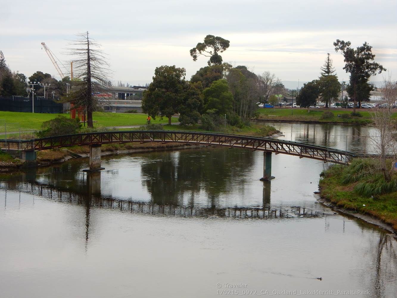 A pedestrian bridge spans calm water, surrounded by trees under a cloudy sky. - Lakeshore Park and Lake Merritt Channel loop