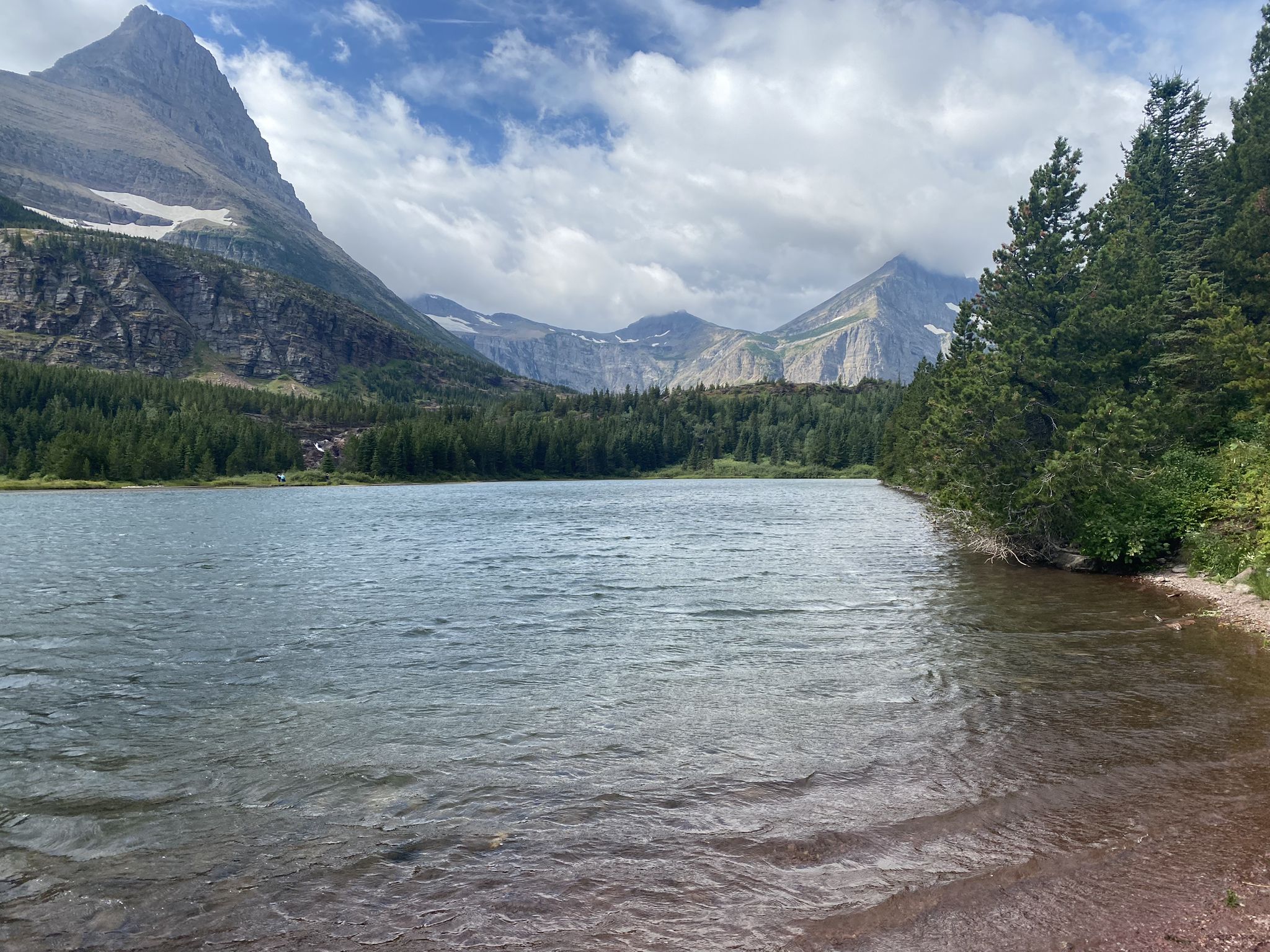 Lake with forested shore and distant mountains under cloudy sky. - Redrock Lake loop from Many Glacier Lodge