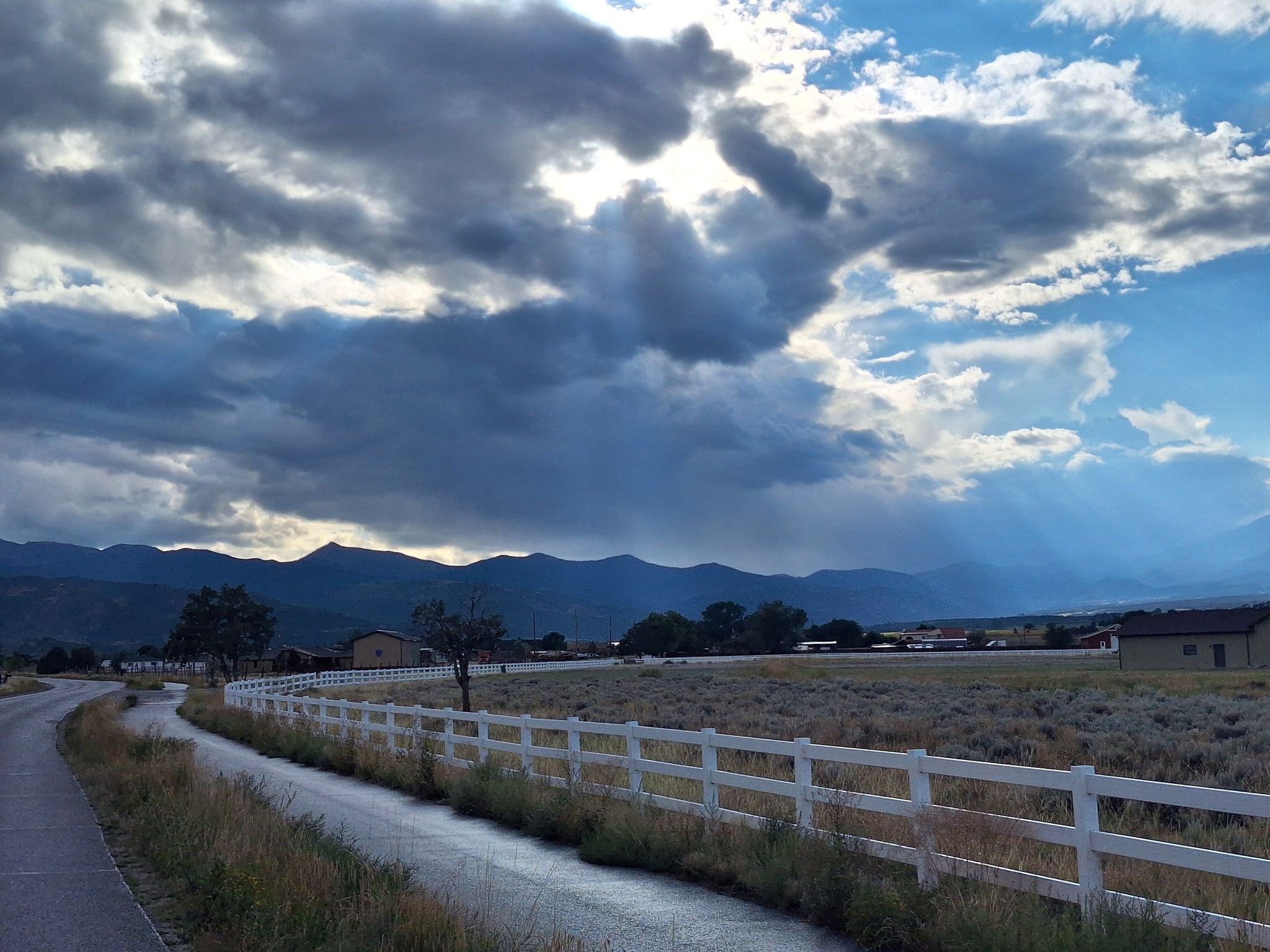 A road and white fence under a dramatic cloudy sky with sunbeams over mountains. - Poncha Springs – The Salida Museum loop from Poncha Springs