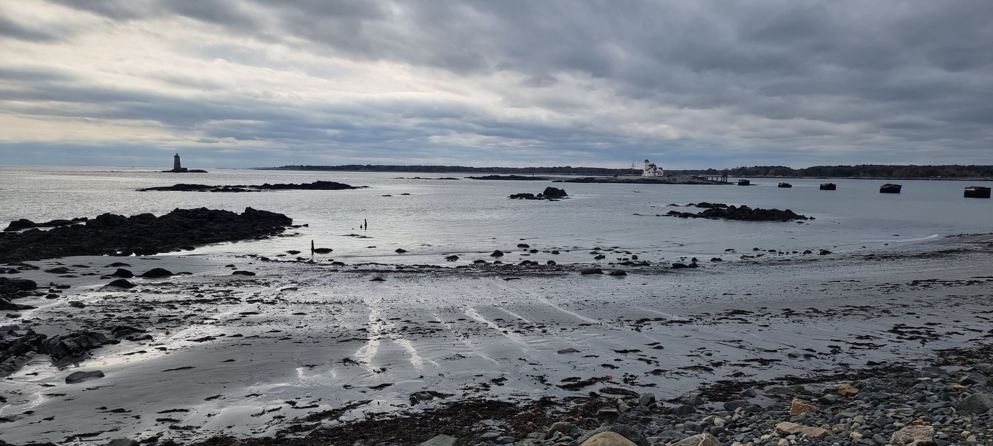 Overcast beach scene with sand, rocks, calm ocean, and distant lighthouse. - Fort Foster Loop Trail