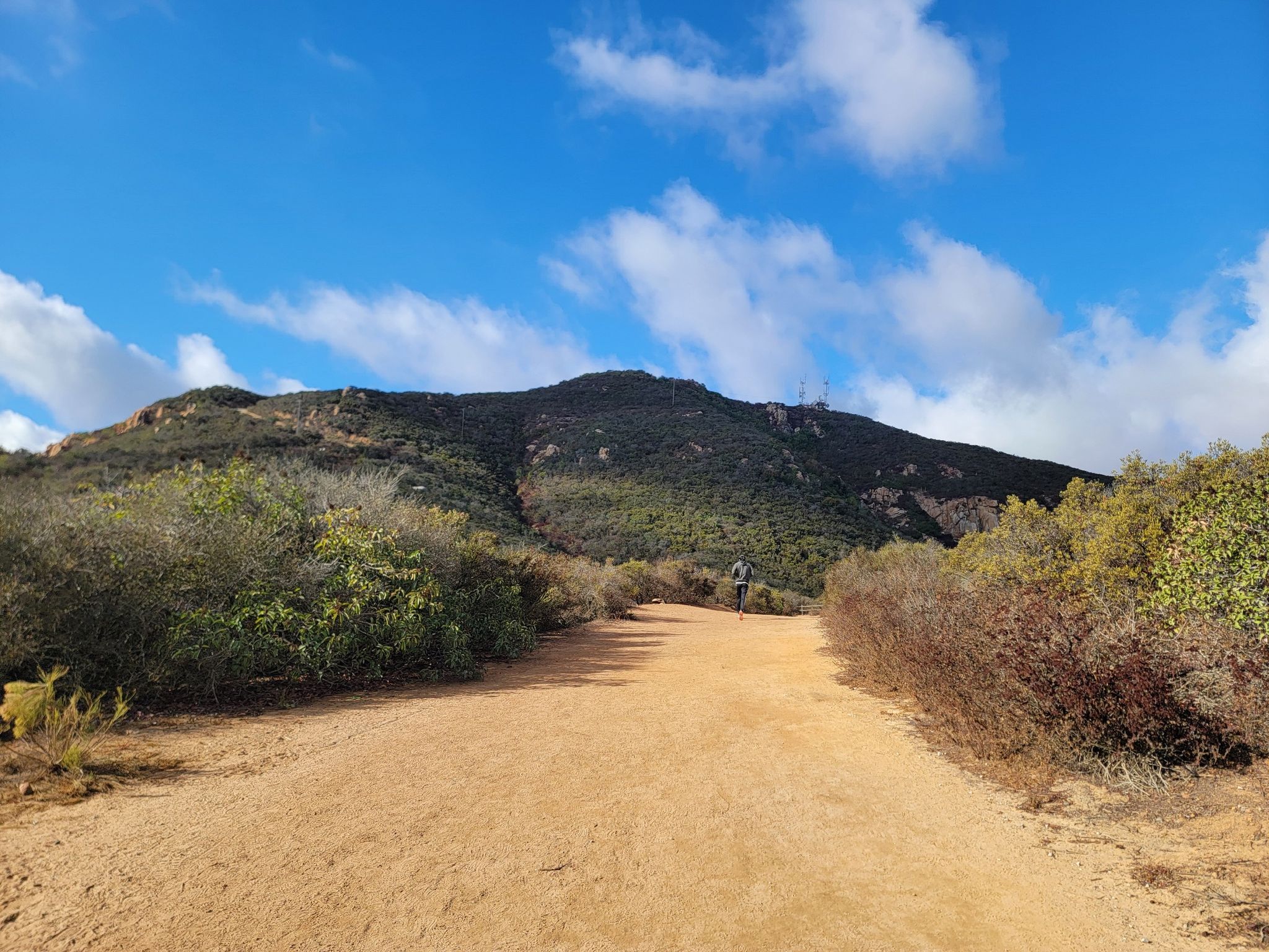 Person walking on a wide dirt road towards a large green mountain. - View from Cowles Mountain loop from San Carlos