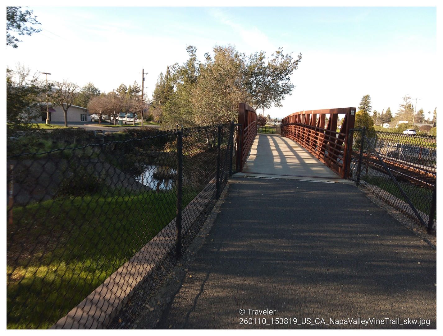 A paved path leading to a metal pedestrian bridge over water on a sunny day. - Napa Valley and Dry Creek Loop