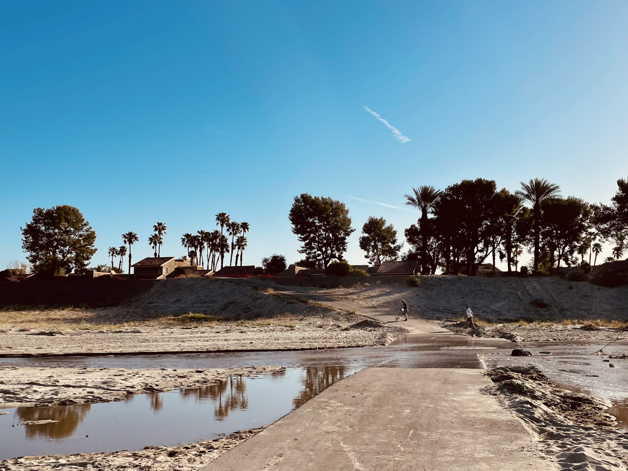 Concrete path crosses shallow water towards sandy banks with distant trees and figures. - Butler Abrams Trail