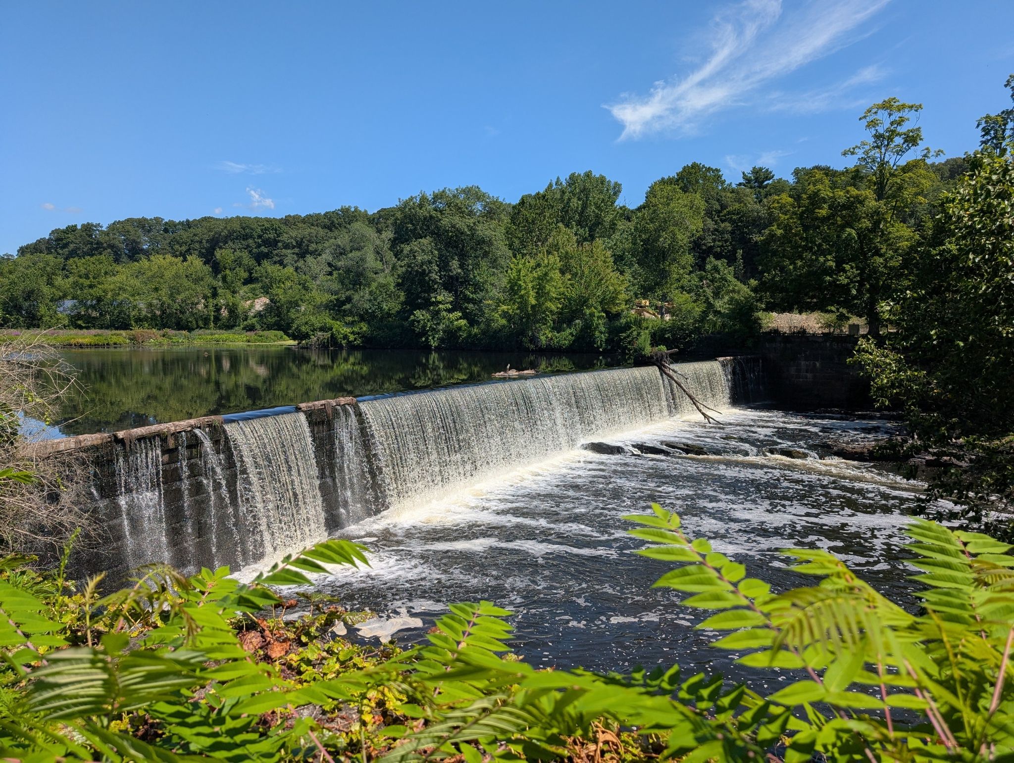 A waterfall cascades over a dam, surrounded by green trees under a blue sky. - Along the Blackstone River – Blackstone River Bikeway loop from Lincoln