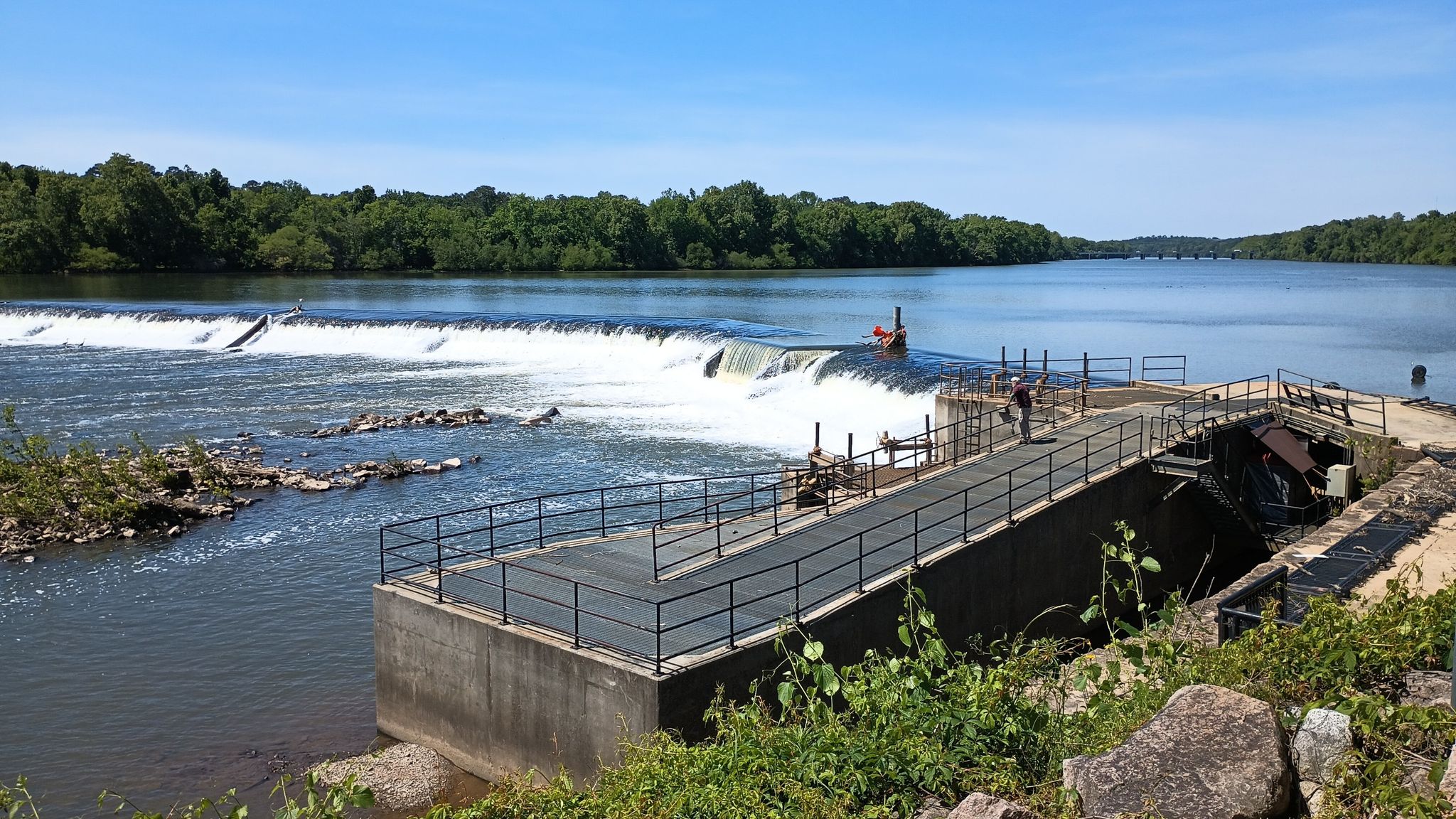 Wide river with a dam, concrete structure, and lush green trees under bright sun. - Three Rivers Greenway