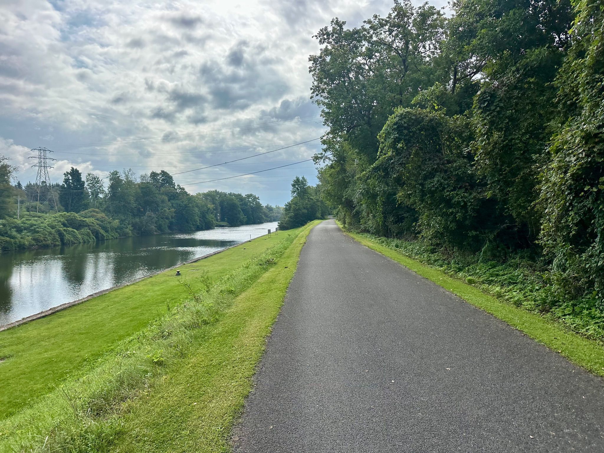 Paved path alongside a wide river with green banks and distant power lines. - Utica Marsh – Erie Canalway Trail loop from Canalway Trail