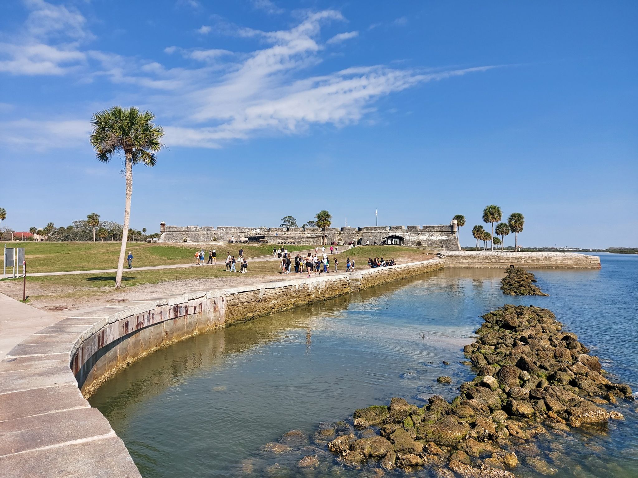 People walk near a stone fort wall by the water on a sunny day. - Castillo de San Marcos – Cathedral loop from Saint Augustine