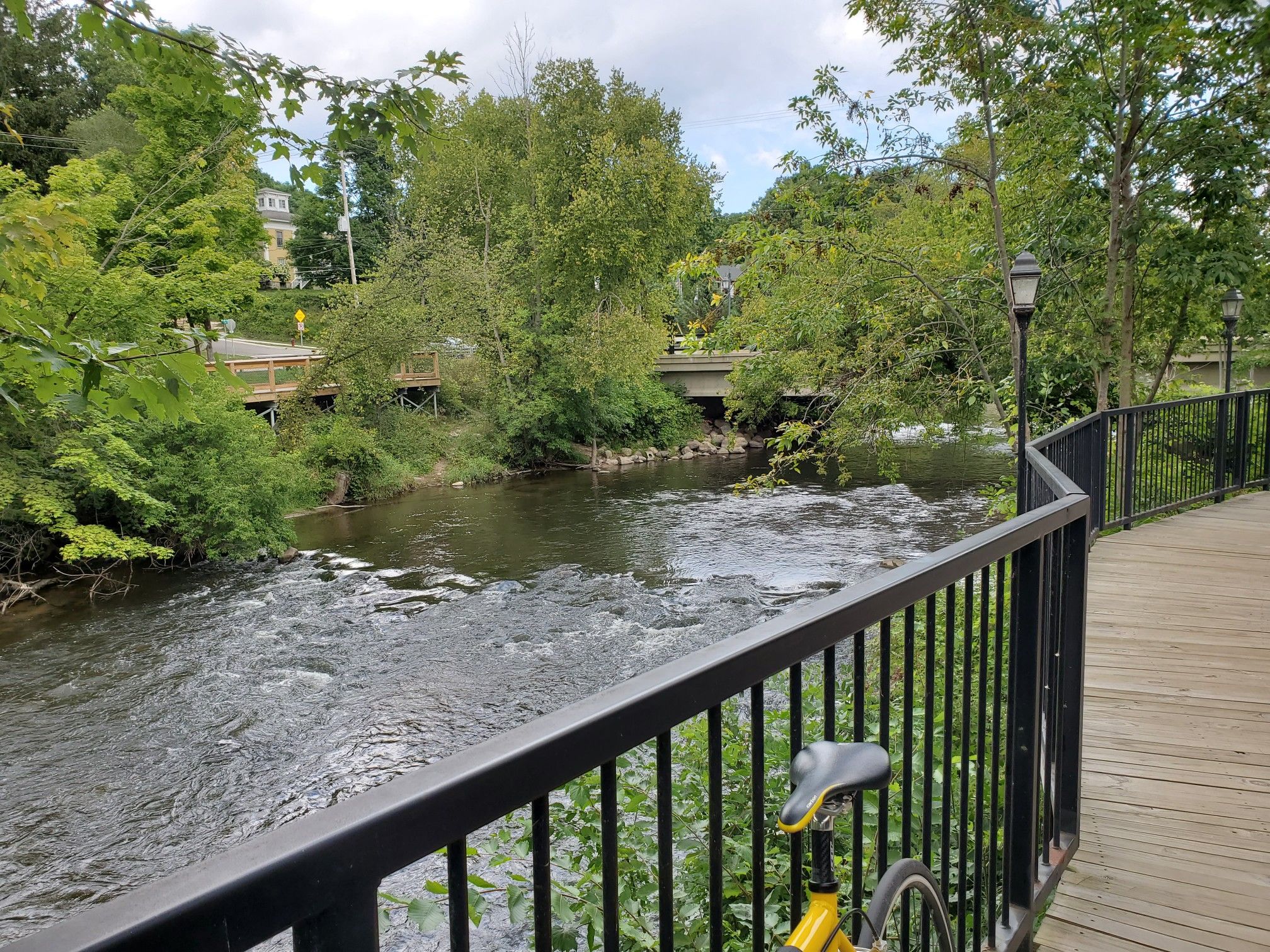 A river flows past a wooden boardwalk with a bicycle, surrounded by green trees. - Fred Meijer White Pine Trail