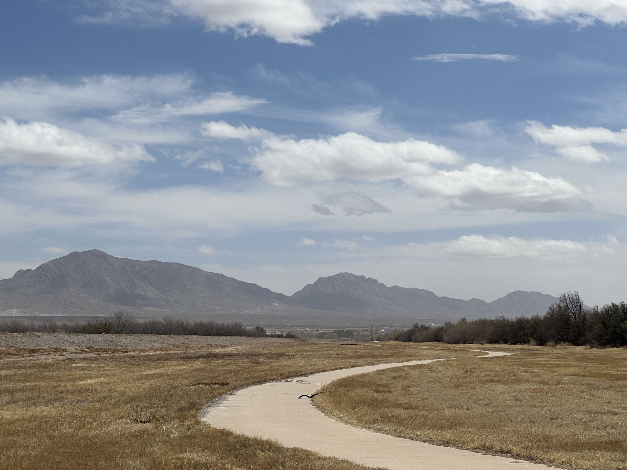 A winding path through dry grass leads towards distant mountains under a cloudy sky. - River Park Trail